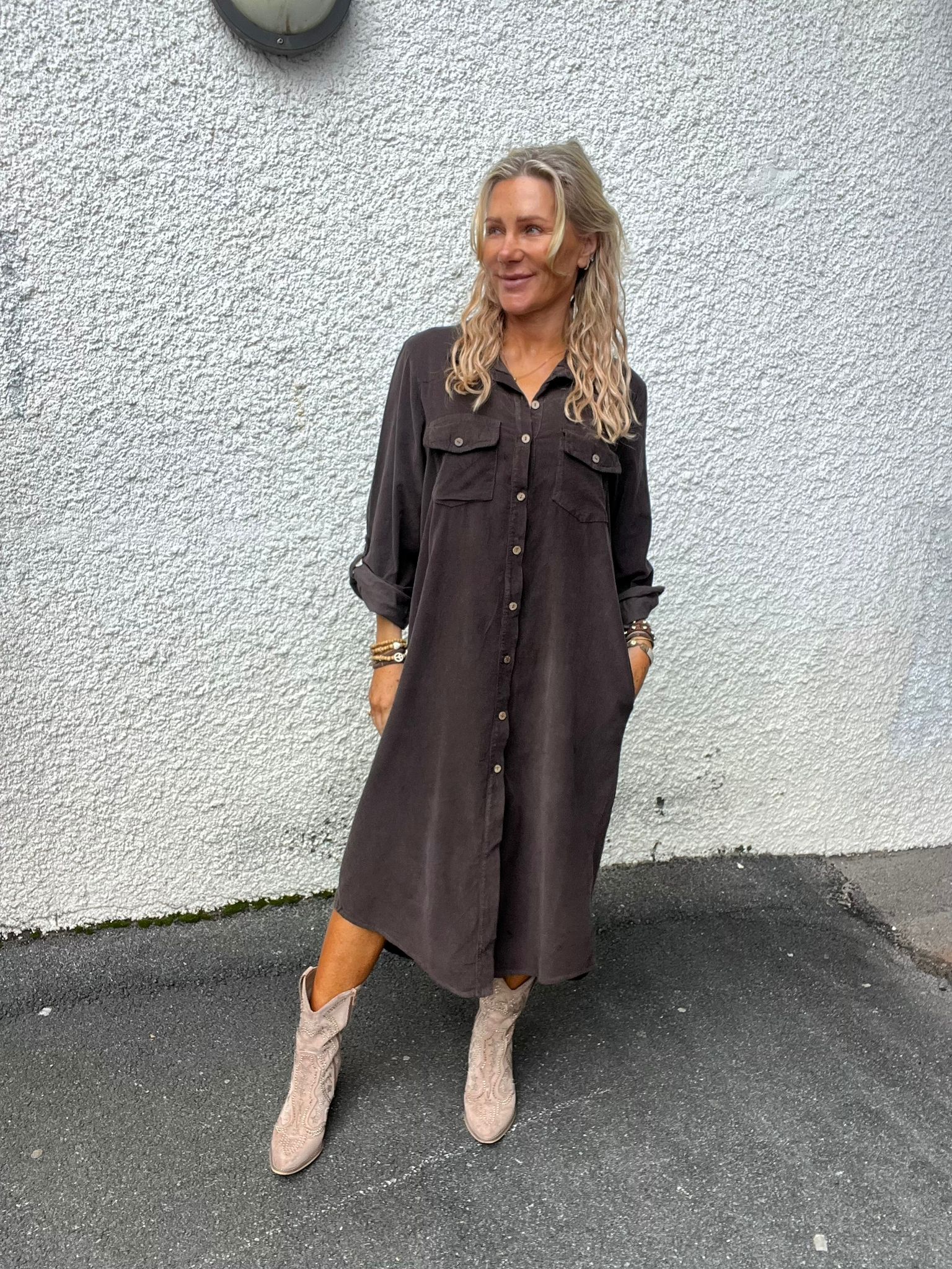 Kvinna med långt blont hår står och ler med händerna i fickorna, klädd i mörk skjortklänning och ljusa cowboyboots. Hon lutar sig mot en vit putsad vägg utomhus, med en vägglampa ovanför.