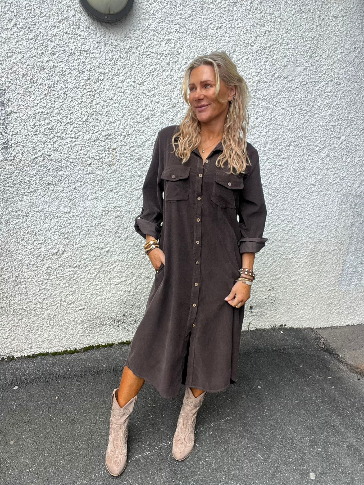 Leende kvinna med långt blont hår står lutad mot en vit putsad vägg, med händerna i fickorna. Hon bär en mörk skjortklänning och ljusa mönstrade boots på asfalt, med armband på handlederna.