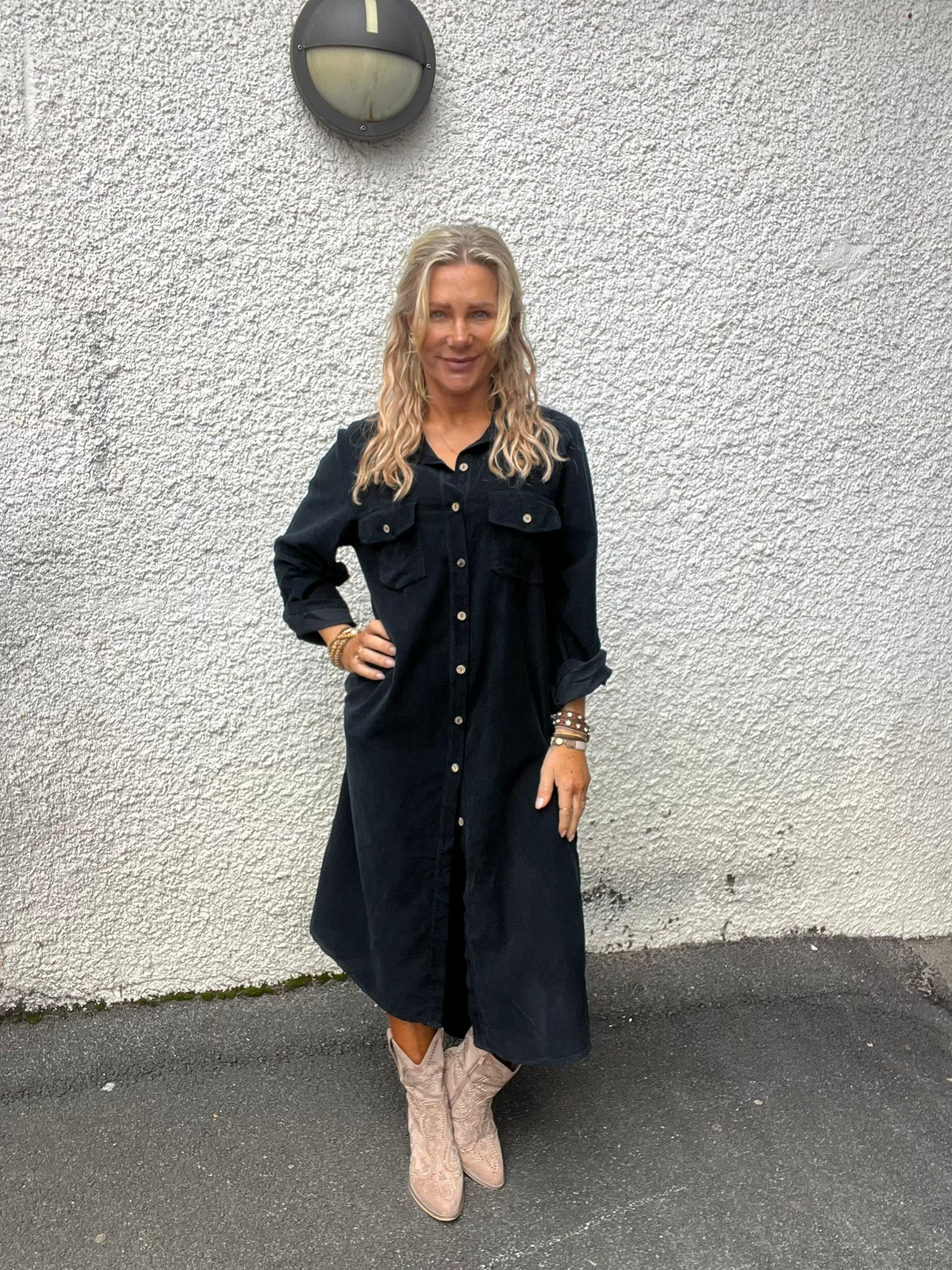 Kvinna med långt blont hår står och ler med ena handen på höften framför en vit, grovputsad vägg. Hon bär en svart skjortklänning och ljusa cowboyboots, med en rund vägglampa ovanför.