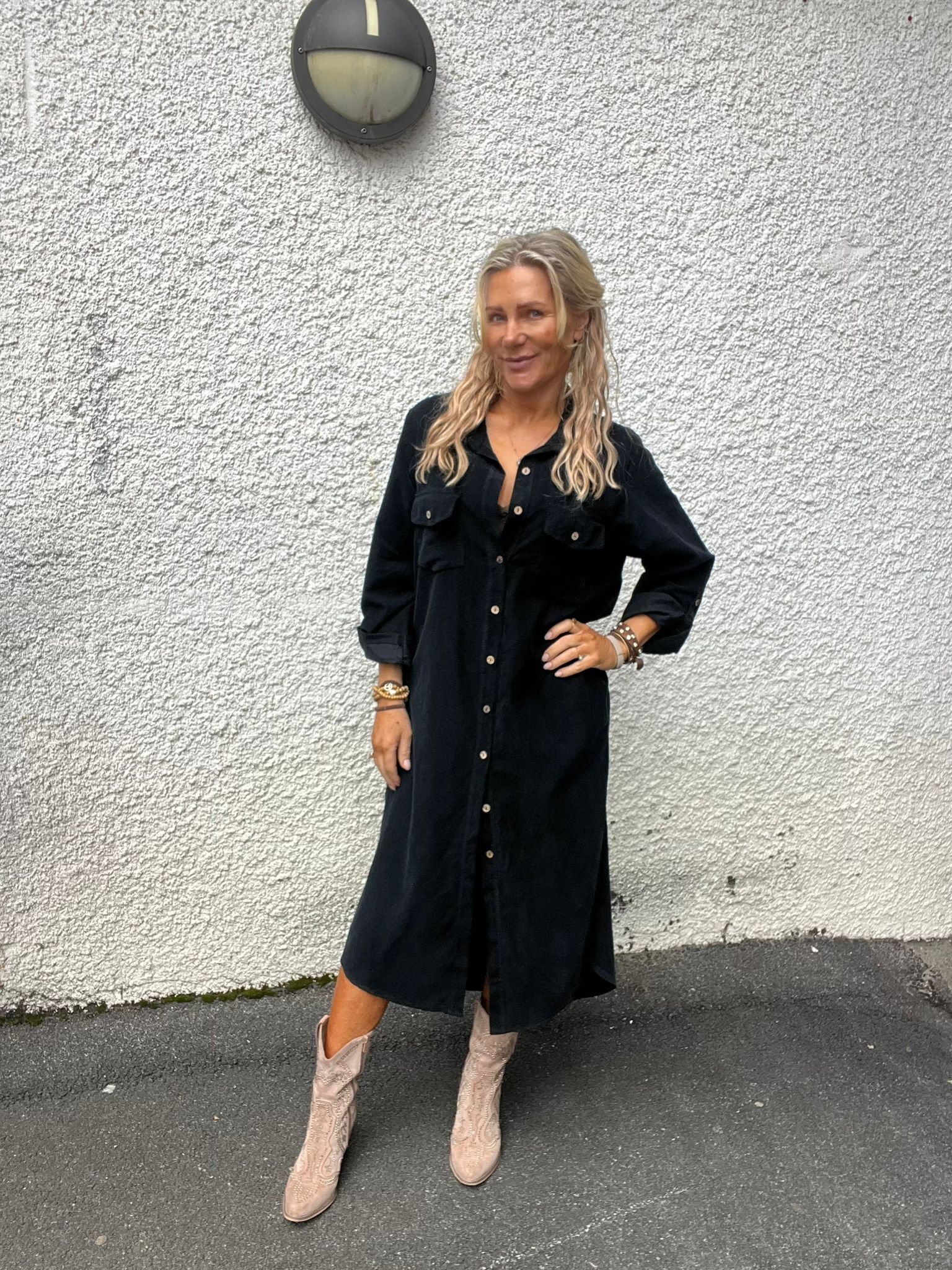 Kvinna med långt blont hår poserar med ena handen på höften framför en vit, grovputsad vägg med rund vägglampa ovanför. Hon bär svart skjortklänning och ljusa cowboyboots samt armband.