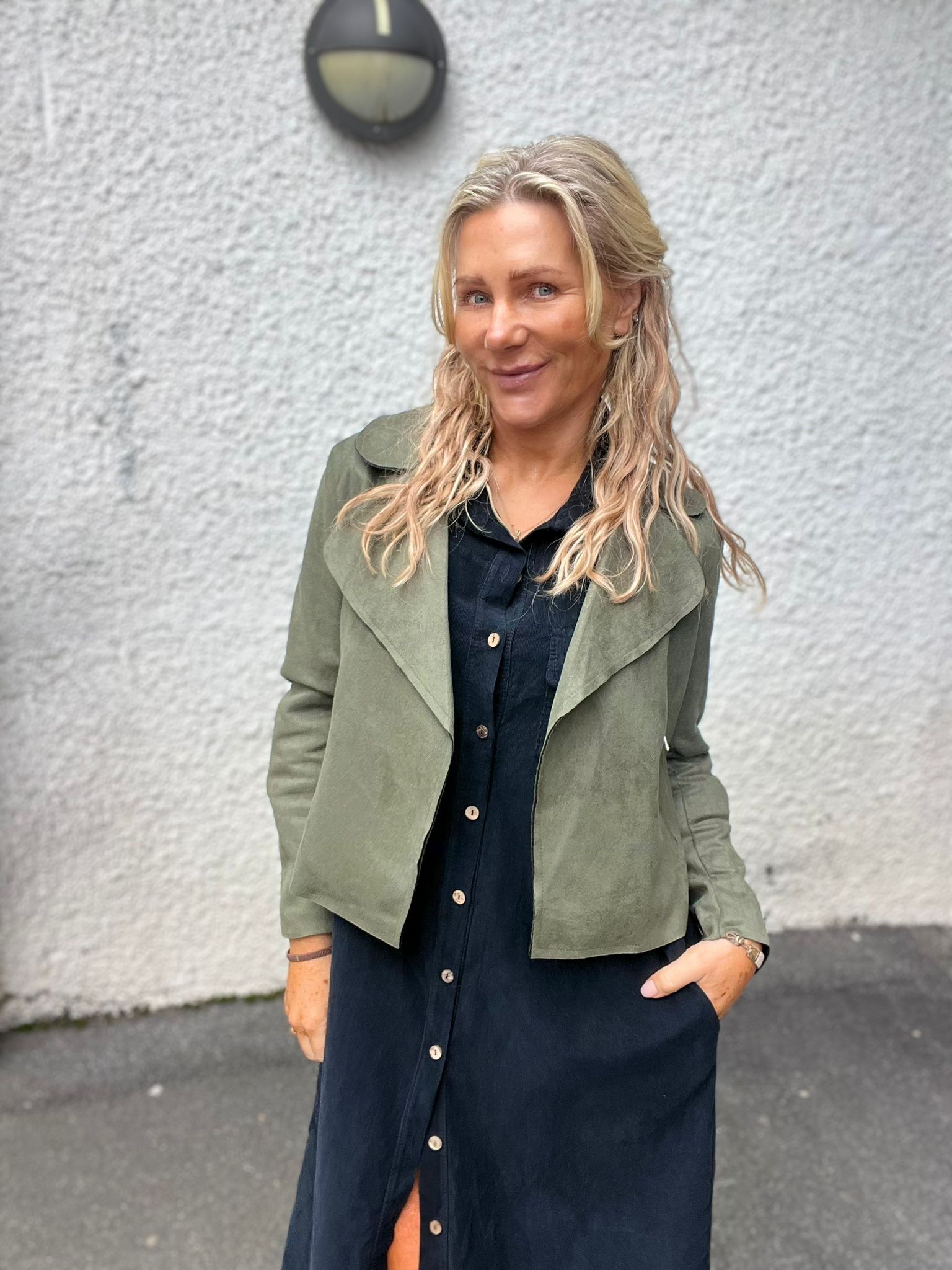 Blond kvinna står och ler mot kameran med händerna i fickorna, klädd i mörk knappad klänning och olivgrön jacka. Hon står utomhus framför en ljus, grovputsad vägg med en rund vägglampa ovanför.