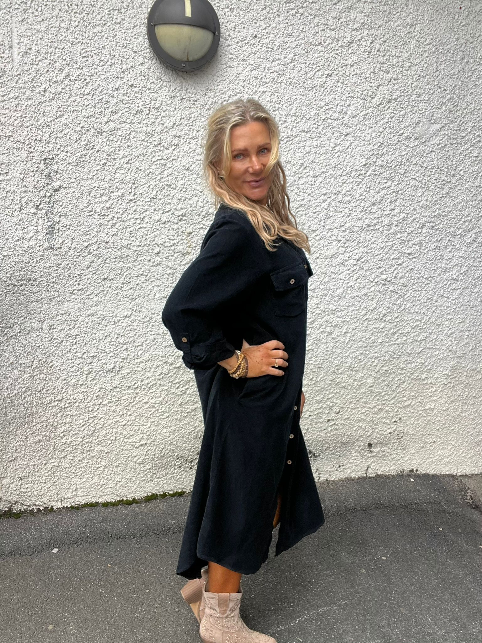 Blond kvinna poserar i profil med handen på höften och ler mot kameran. Hon bär en svart skjortklänning och ljusa cowboyboots, står på asfalt framför en vit, grovputsad vägg med en rund vägglampa ovanför.