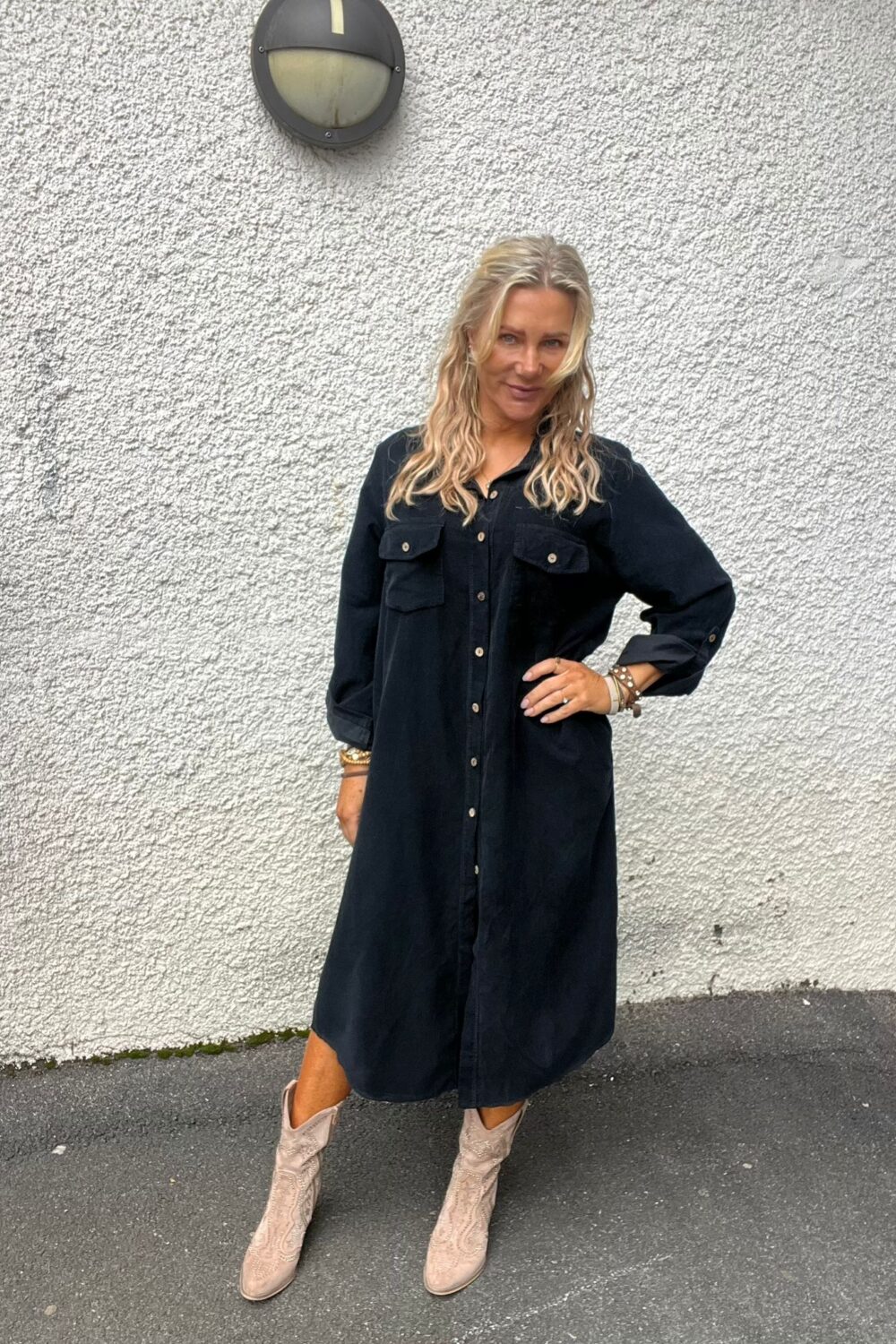 Kvinna med långt blont hår poserar med ena handen på höften och ler lätt mot kameran. Hon bär svart skjortklänning och ljusa boots, står på asfalt framför en vit, grovputsad vägg med en rund vägglampa.