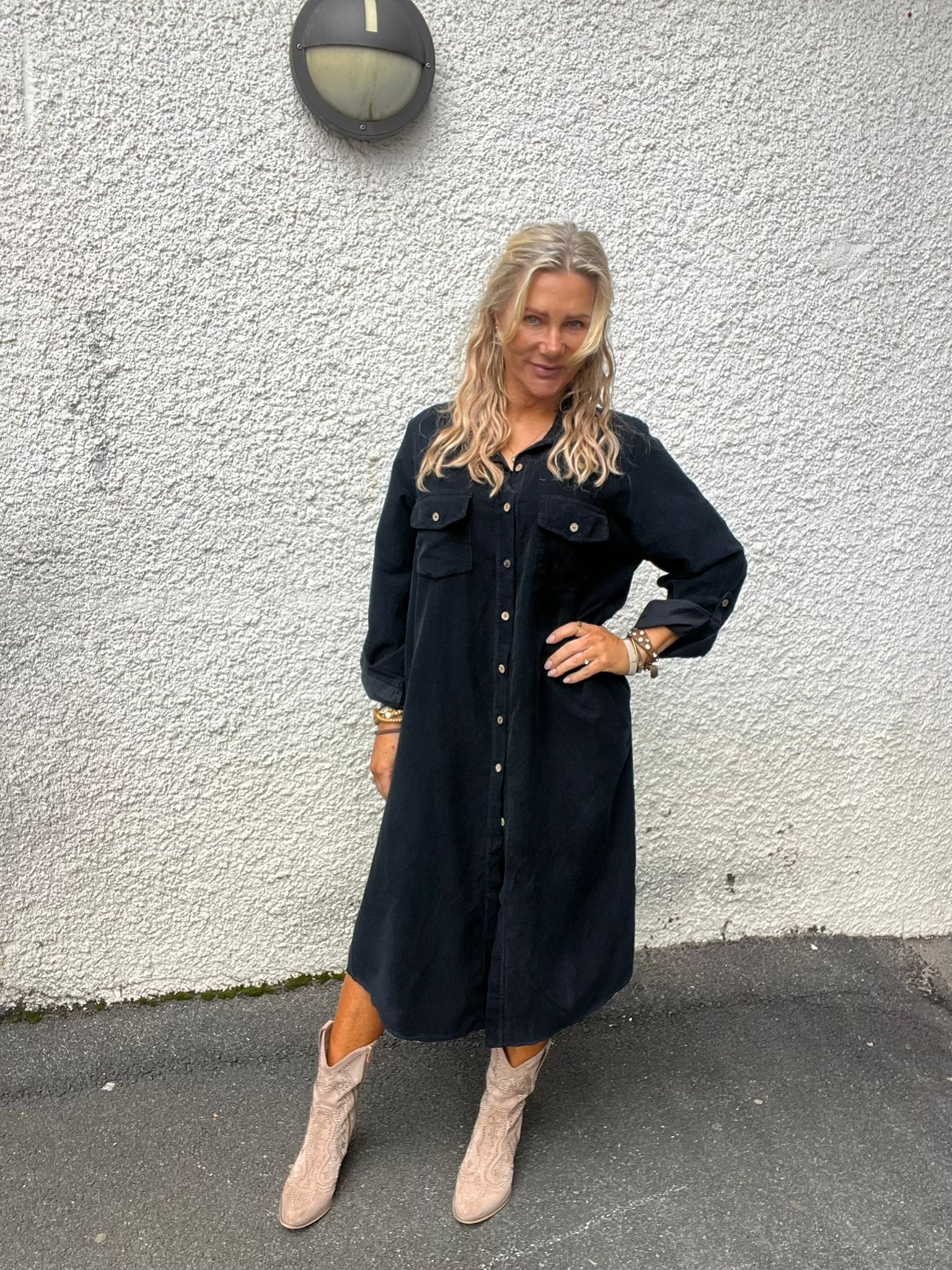 Kvinna med långt blont hår poserar med ena handen på höften och ler lätt mot kameran. Hon bär svart skjortklänning och ljusa boots, står på asfalt framför en vit, grovputsad vägg med en rund vägglampa.