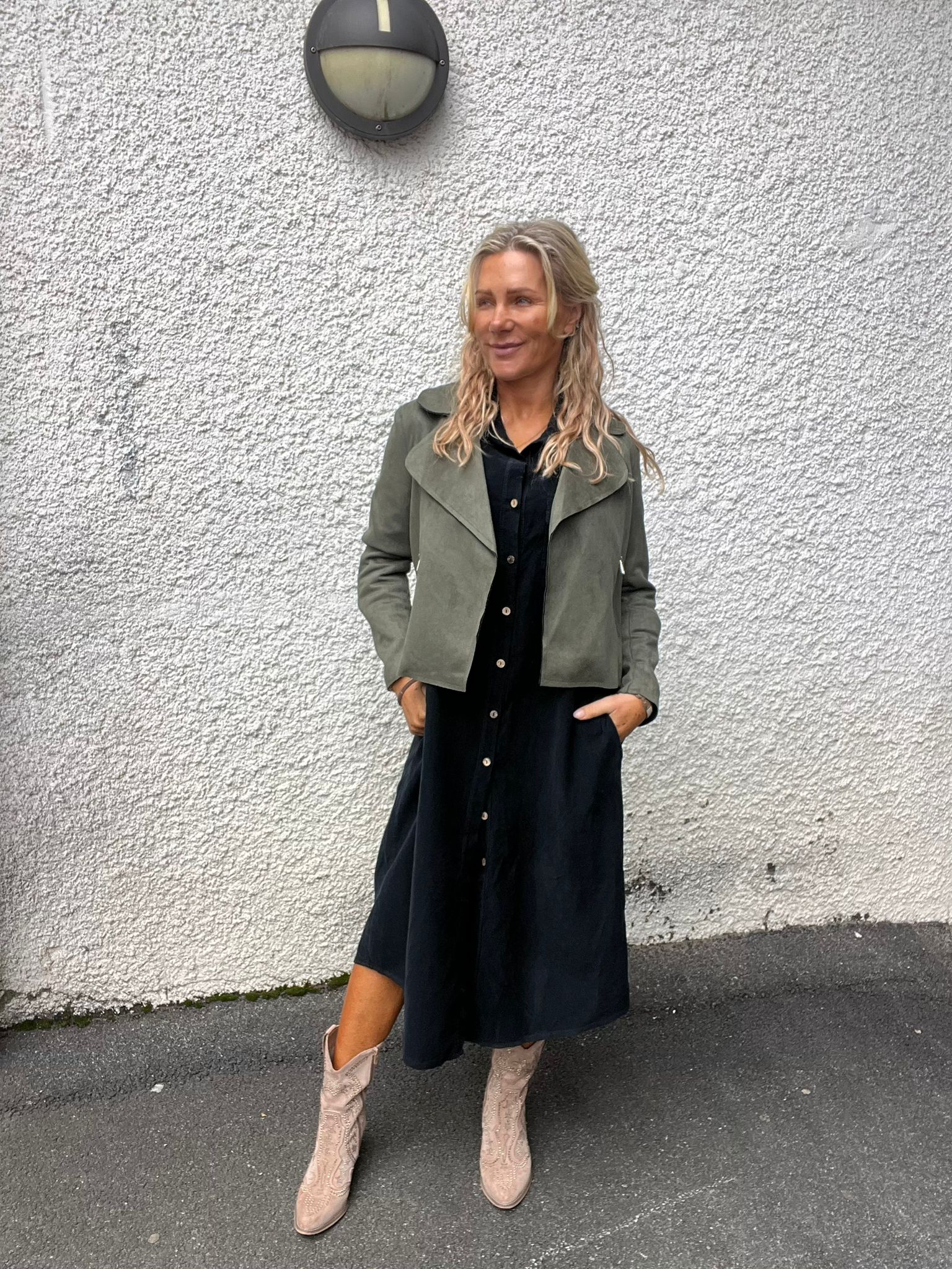 Kvinna står avslappnat med händerna i fickorna och tittar åt sidan, klädd i svart skjortklänning, olivgrön jacka och ljusa boots. Hon poserar på asfalt framför en vit grovputsad vägg med rund vägglampa ovanför.