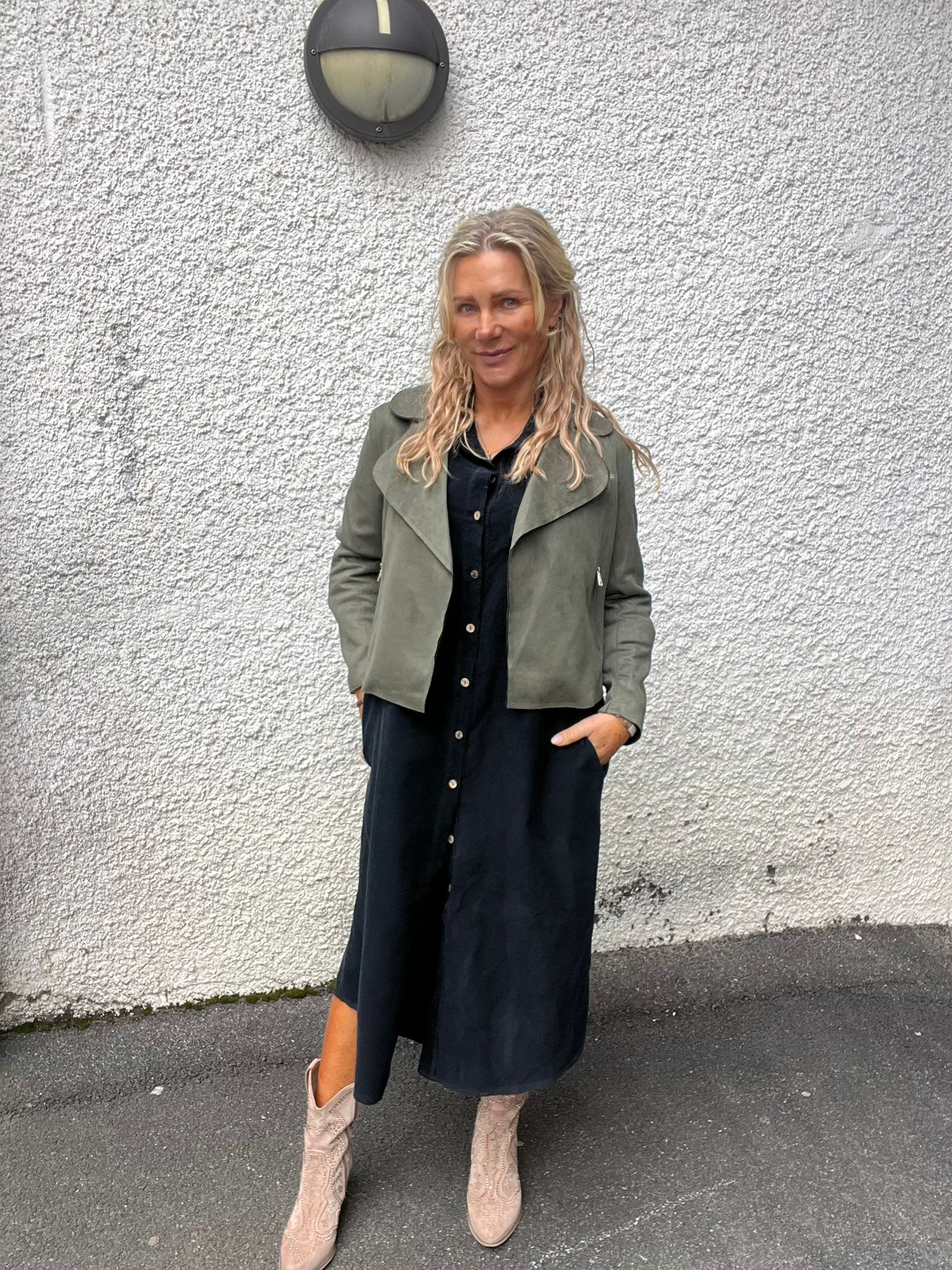 Kvinna med långt blont hår står med händerna i fickorna och ler mot kameran, klädd i svart skjortklänning och grön jacka med ljusa cowboyboots, framför en vit putsad vägg med rund vägglampa.