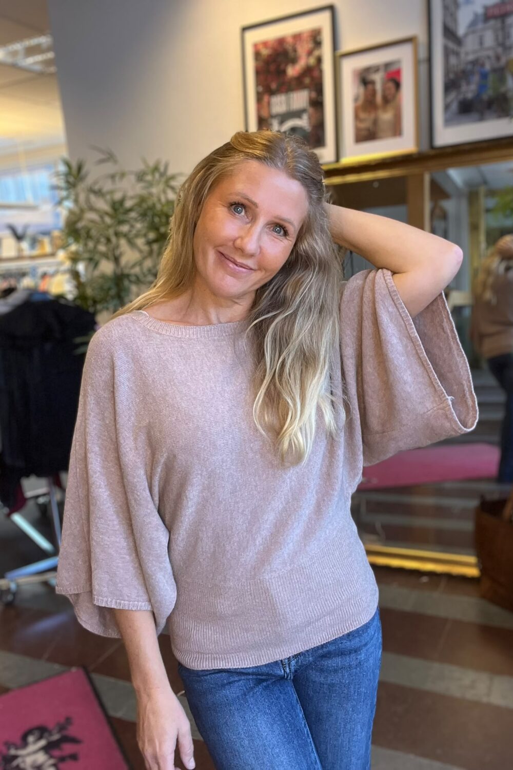 Leende kvinna med långt blont hår står avslappnat med ena handen bakom huvudet i en inomhusmiljö. Hon bär en ljusrosa stickad topp och jeans; bakom syns växter, klädställning och inramade affischer på väggen.