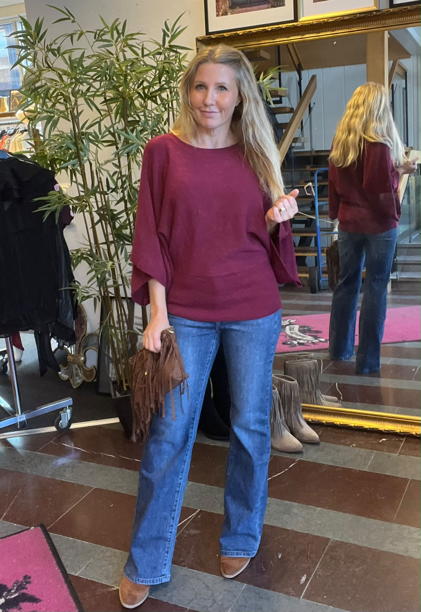 Kvinna med långt blont hår poserar och ler, klädd i vinröd topp med vida ärmar och blå jeans, håller en brun fransväska. Hon står i butiksmiljö framför stor guldramsspegel och grön växt.