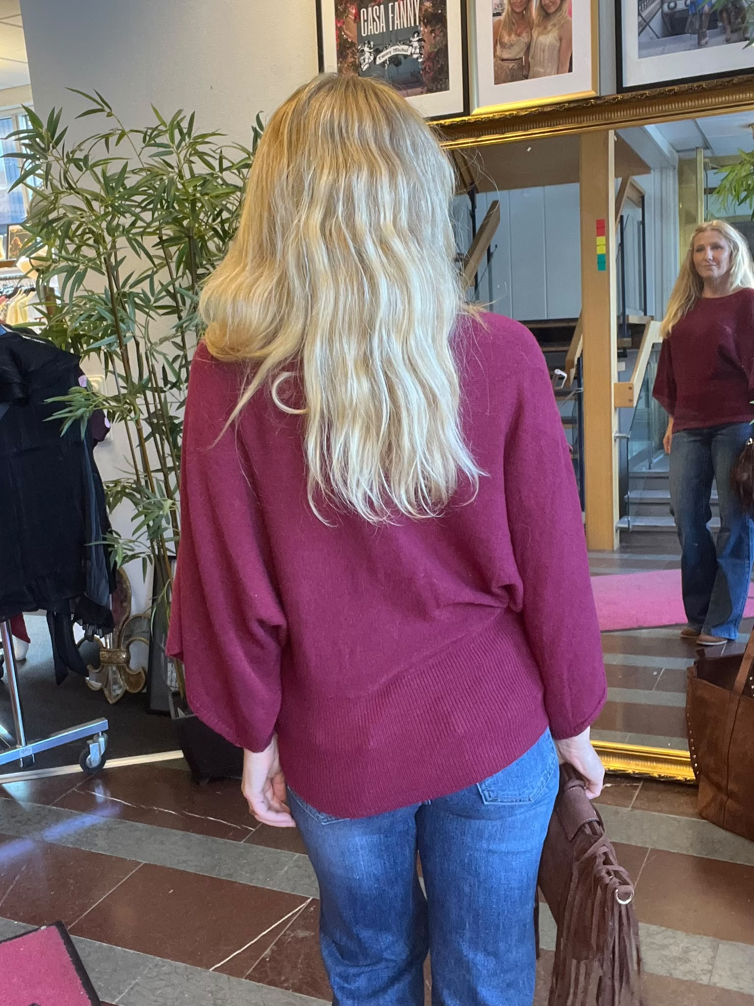 Kvinna med långt blont hår i vinröd tröja står med ryggen mot kameran och tittar i en stor spegel. I spegeln syns hennes ansikte och lokalen med växt, kläder och affischtexten “CASA FANNY”.
