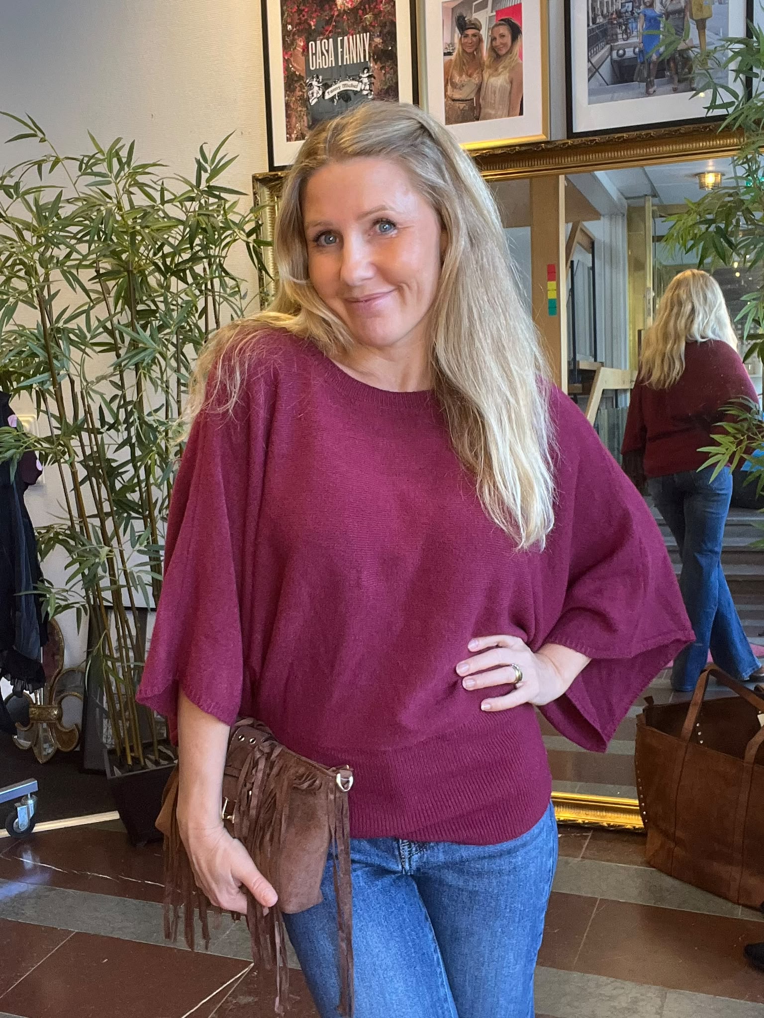 Leende kvinna med långt blont hår poserar med handen på höften och håller en brun fransväska, inomhus bland gröna växter. I bakgrunden syns en spegel och inramade foton; texten “CHISA FANNY”.