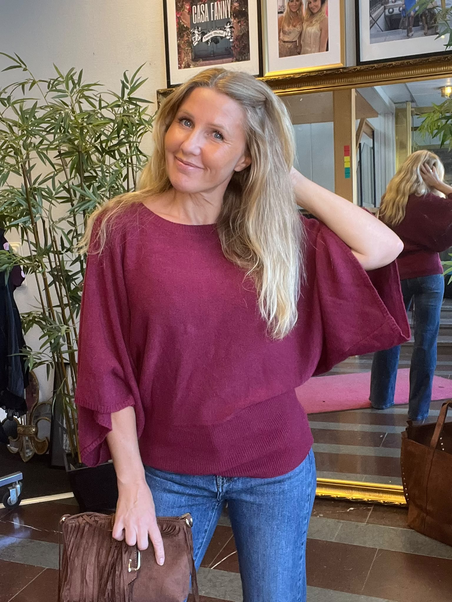 Leende kvinna med långt blont hår poserar med ena handen i nacken och håller en brun väska. Hon står inomhus framför en stor spegel, växter och inramade foton; texten “CASA FANNY” syns.