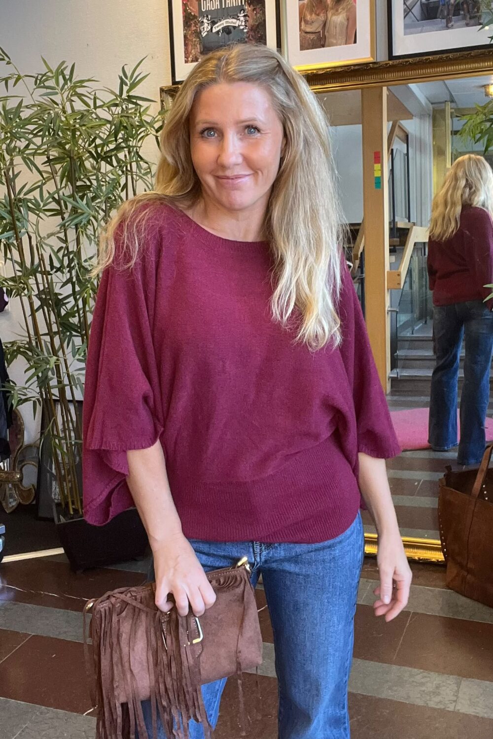 Leende kvinna i vinröd stickad tröja och blå jeans står och håller en brun fransväska inomhus. Bakom henne syns en stor guldramad spegel som visar hennes rygg, växter och tavlor på väggen.