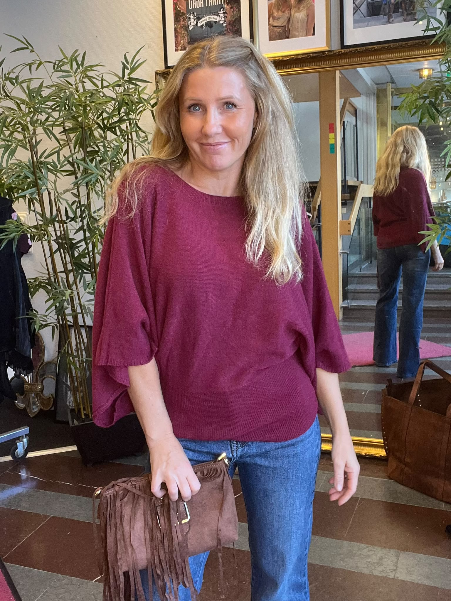 Leende kvinna i vinröd stickad tröja och blå jeans står och håller en brun fransväska inomhus. Bakom henne syns en stor guldramad spegel som visar hennes rygg, växter och tavlor på väggen.