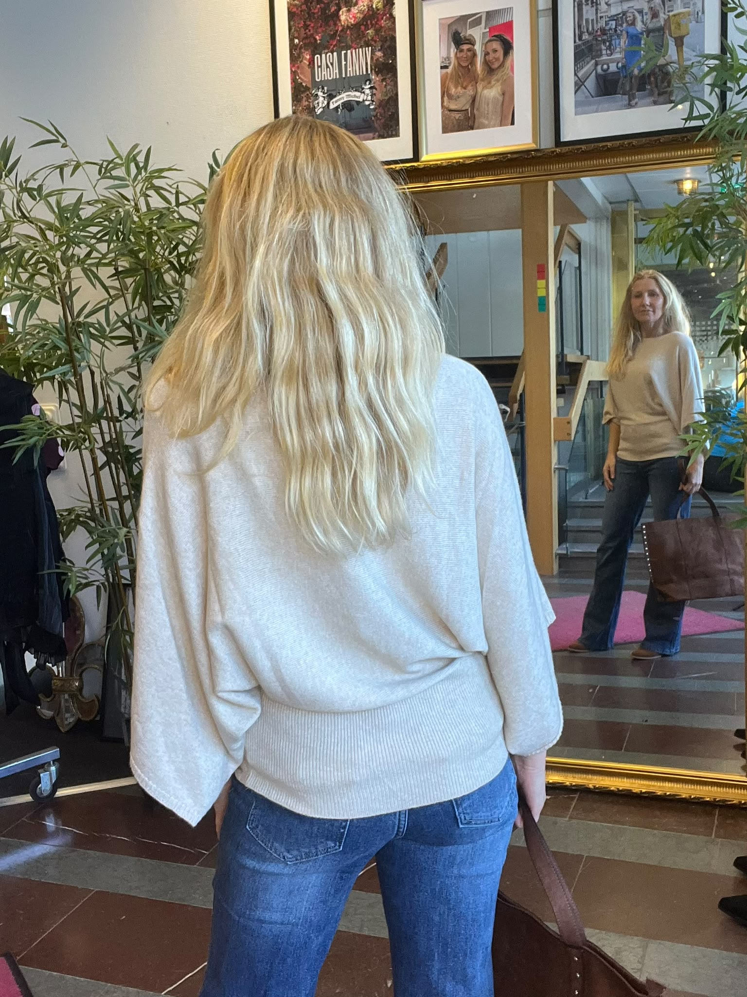 Blond kvinna står med ryggen mot kameran och tittar på sin spegelbild i en stor guldramsspegel i en lobby med växter. Ovanför hänger ramar; en affisch med texten “CASA FANNY”.