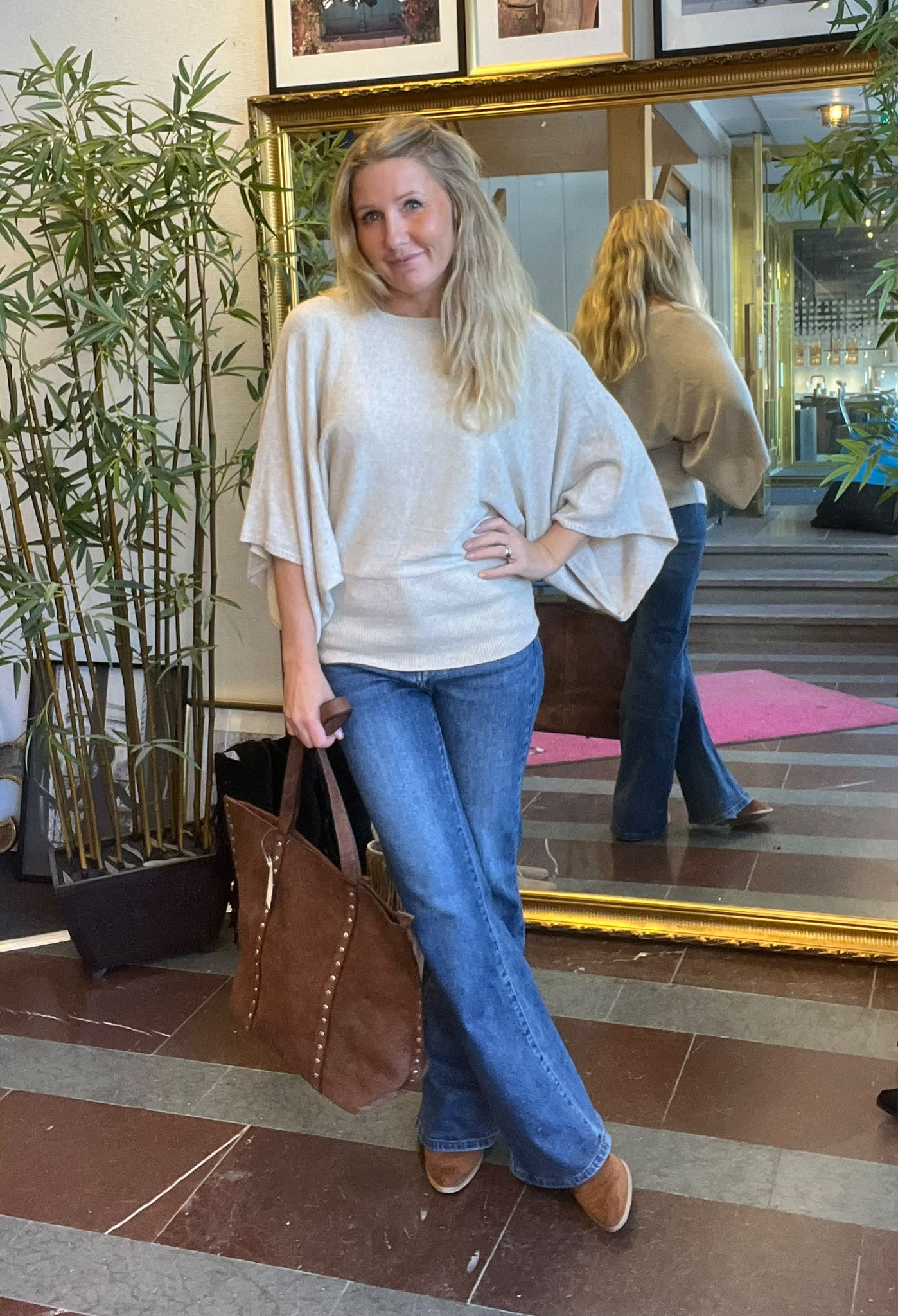Kvinna med långt blont hår poserar med handen på höften och håller en brun läderväska. Hon bär ljus, vidärmad tröja och blå jeans, står vid en stor guldramsspegel och en grön växt i en lobby.