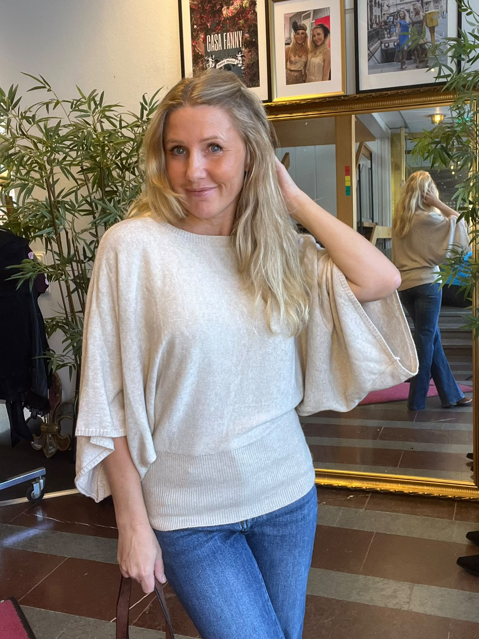 Blond kvinna står inomhus och ler mot kameran, med ena handen i håret och en väska i den andra. Bakom henne syns en spegel, gröna växter och inramade foton med texten “CASA FANNY”.