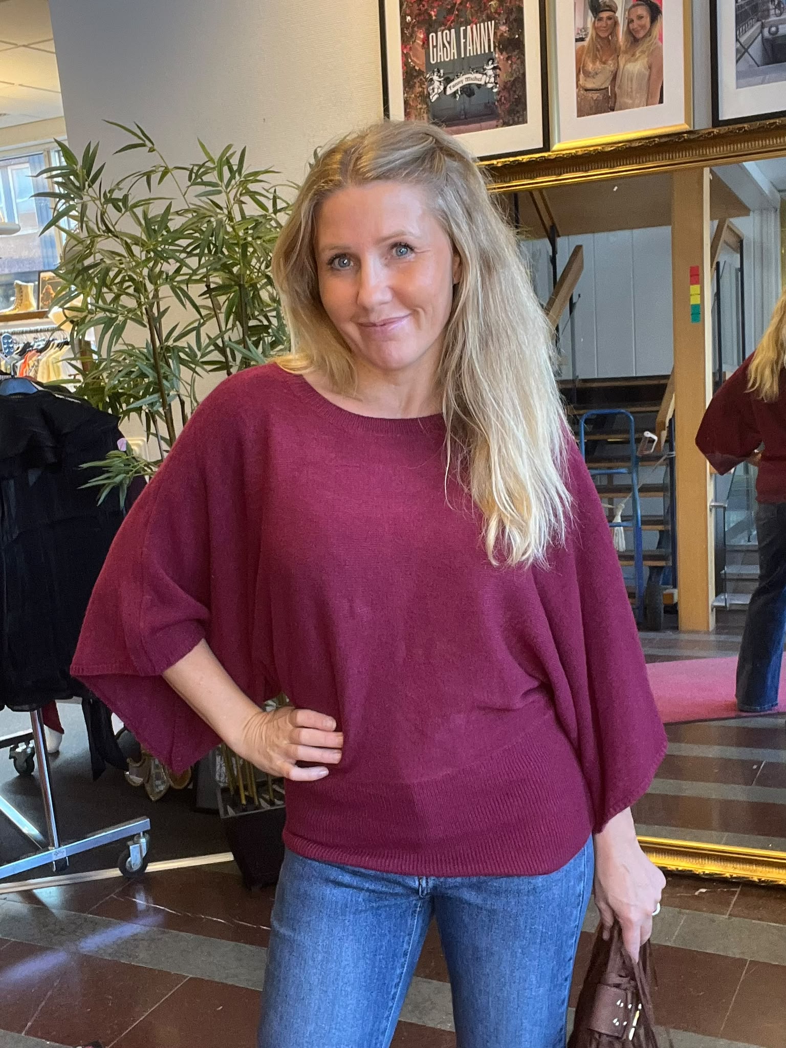 Blond kvinna i vinröd tröja och jeans står och ler med handen på höften i en inomhusmiljö, framför en stor spegel och en grön växt; på väggen syns texten “CASA FANNY”.