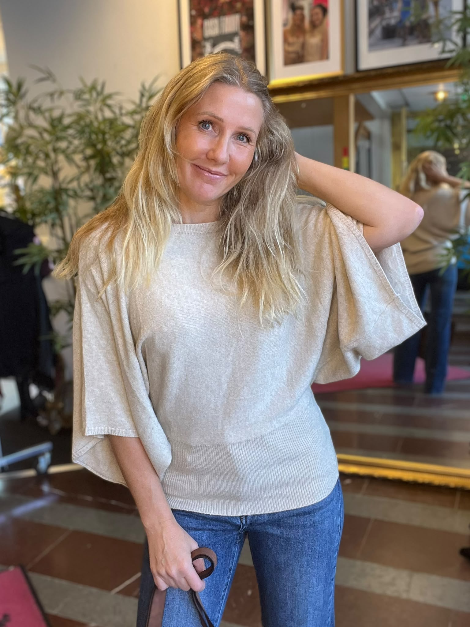 Leende kvinna med långt blont hår poserar och håller ena handen bakom huvudet, klädd i ljus stickad topp och jeans. Hon står inomhus vid en spegel med växter och inramade foton i bakgrunden.