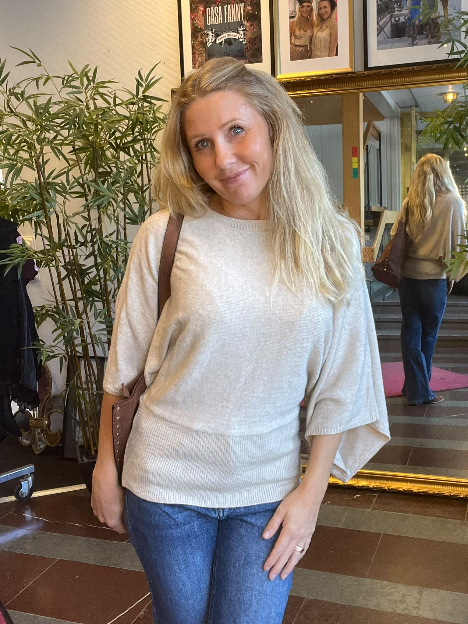 Leende kvinna med långt blont hår står och poserar inomhus med en axelväska, framför en stor guldramsspegel och gröna växter. På väggen syns inramade bilder, inklusive texten “CASA FANNY”.