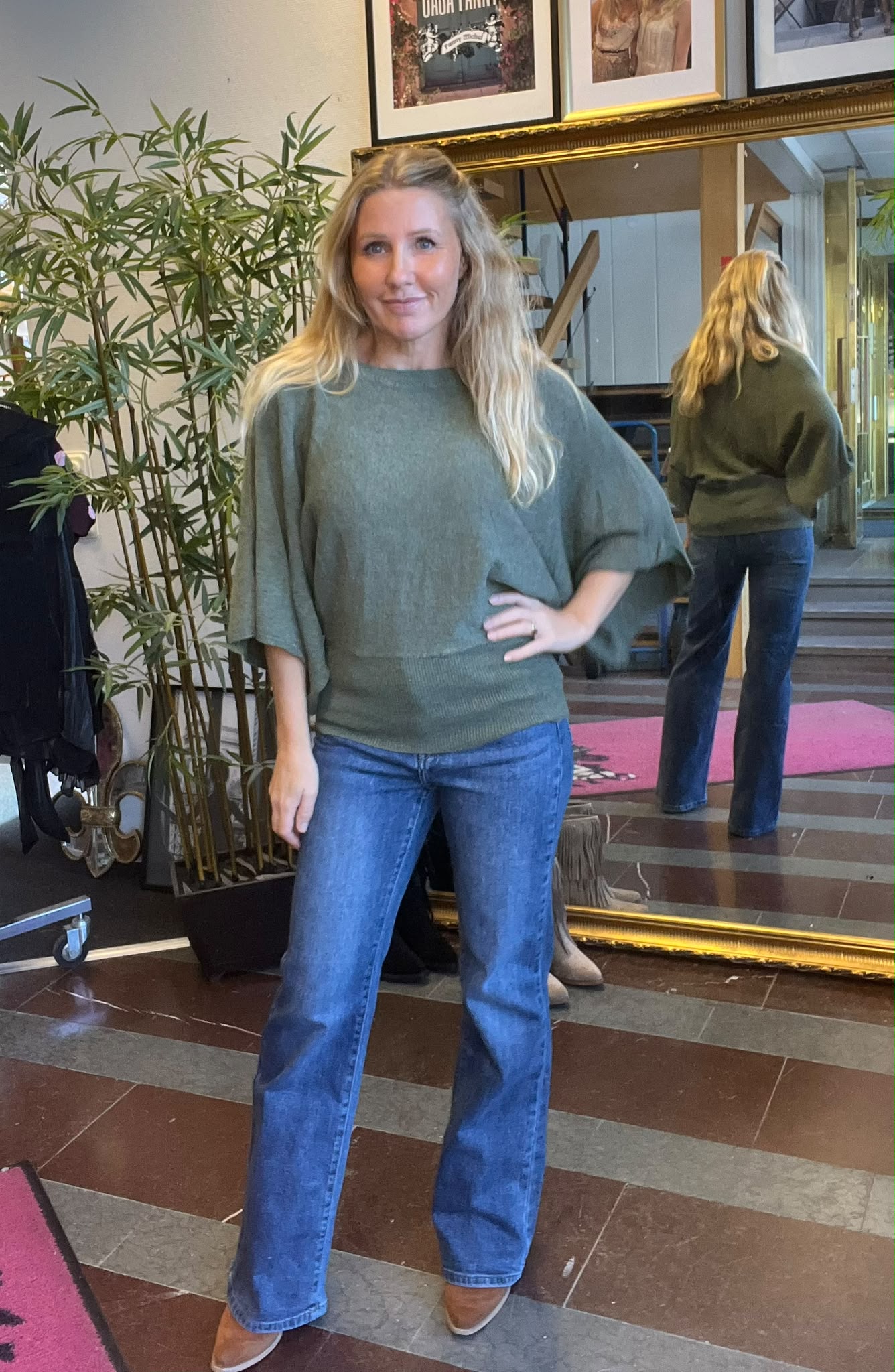 Blond kvinna står och poserar med ena handen på höften, klädd i grön stickad tröja, blå utsvängda jeans och bruna skor. Hon står inomhus framför en stor spegel, med växt och tavlor i bakgrunden.