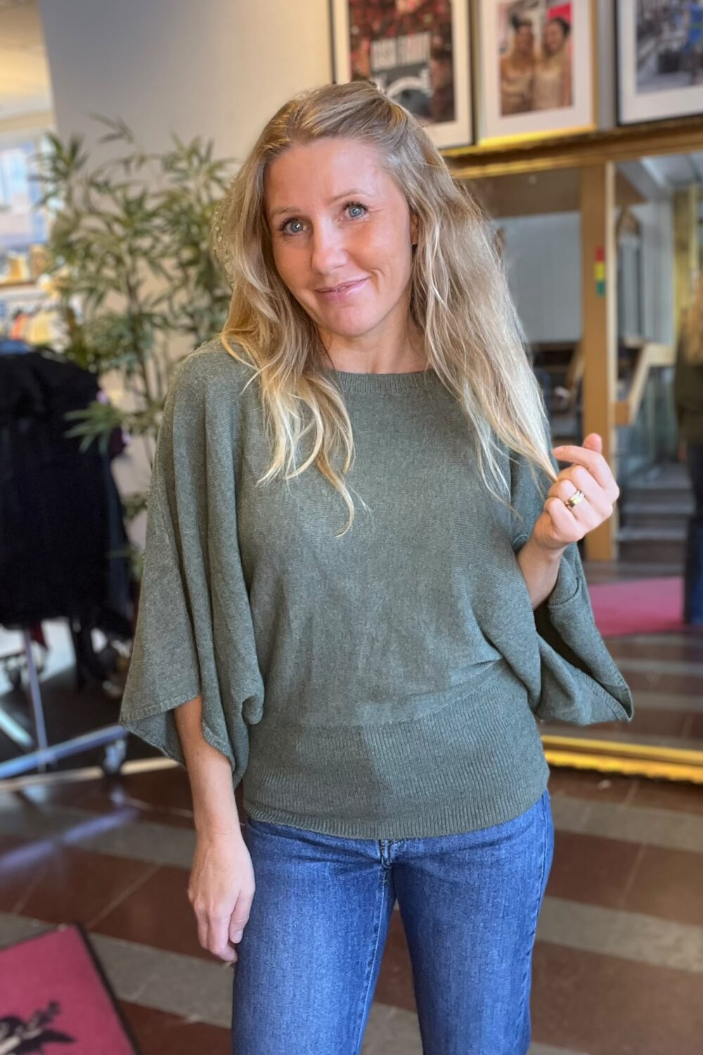 Leende kvinna med långt blont hår poserar och håller upp handen med ring, klädd i grön stickad tröja och blå jeans. Hon står inomhus i en salong med spegel, växter och inramade foton.