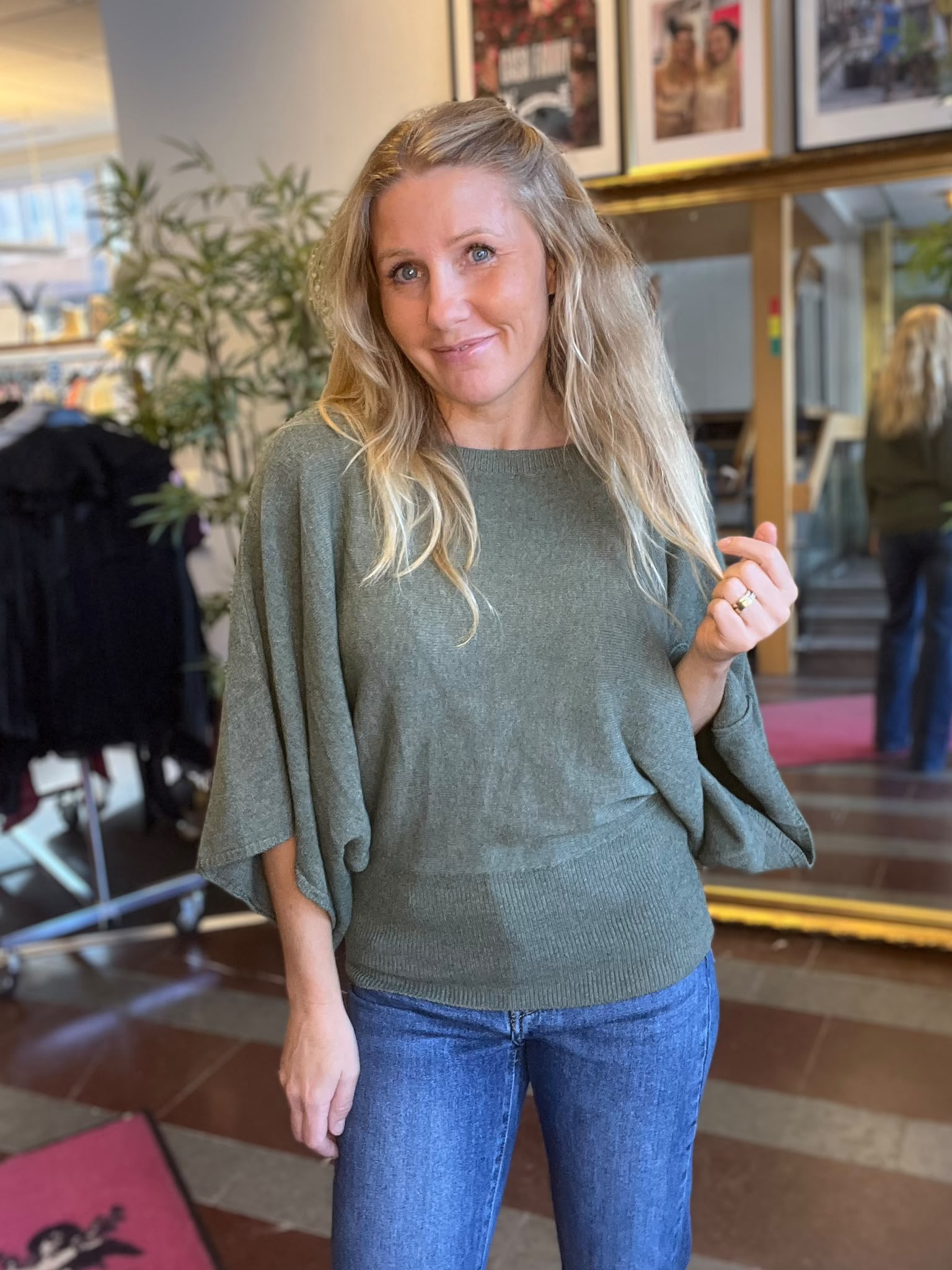 Leende kvinna med långt blont hår poserar och håller upp handen med ring, klädd i grön stickad tröja och blå jeans. Hon står inomhus i en salong med spegel, växter och inramade foton.