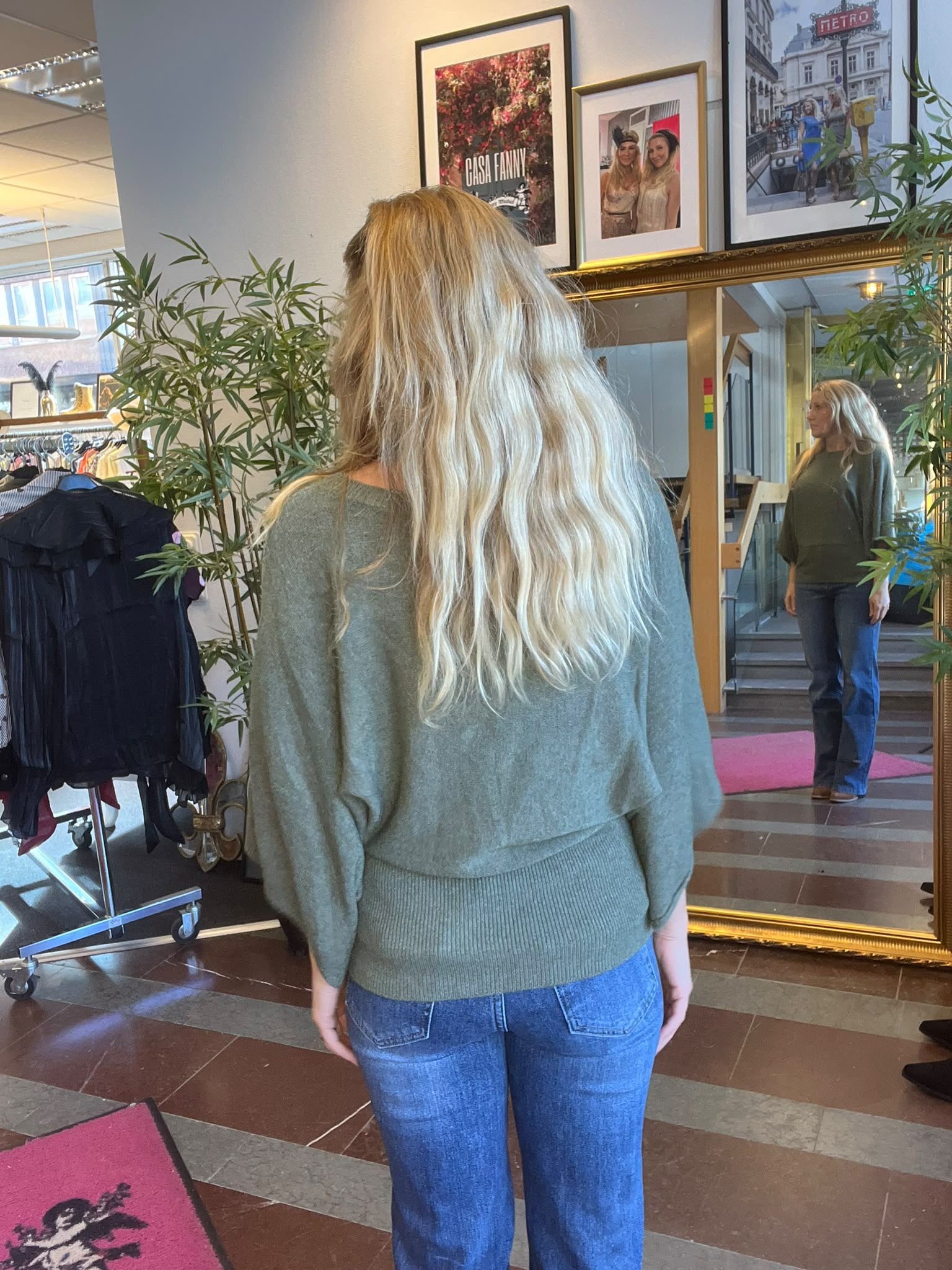 Kvinna med långt blont hår står med ryggen mot kameran och tittar på sin spegelbild i en stor guldramsspegel i en klädbutik, med klädställning och växter runt. Tavla med texten “CASA FANNY”.