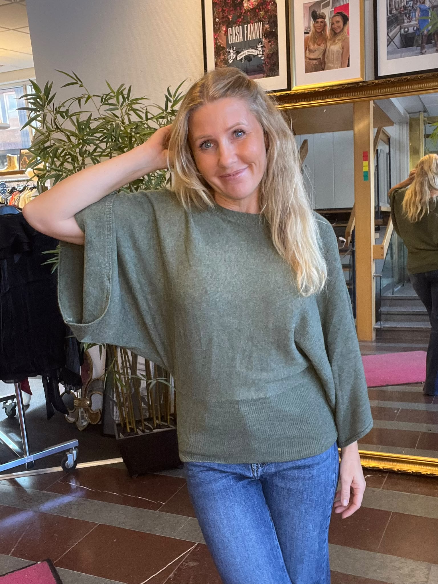 Leende kvinna med långt blont hår står inomhus och håller ena handen bakom huvudet. Hon bär grön stickad tröja och jeans; bakom syns spegel, växt och inramade bilder med texten “CASA FANNY”.