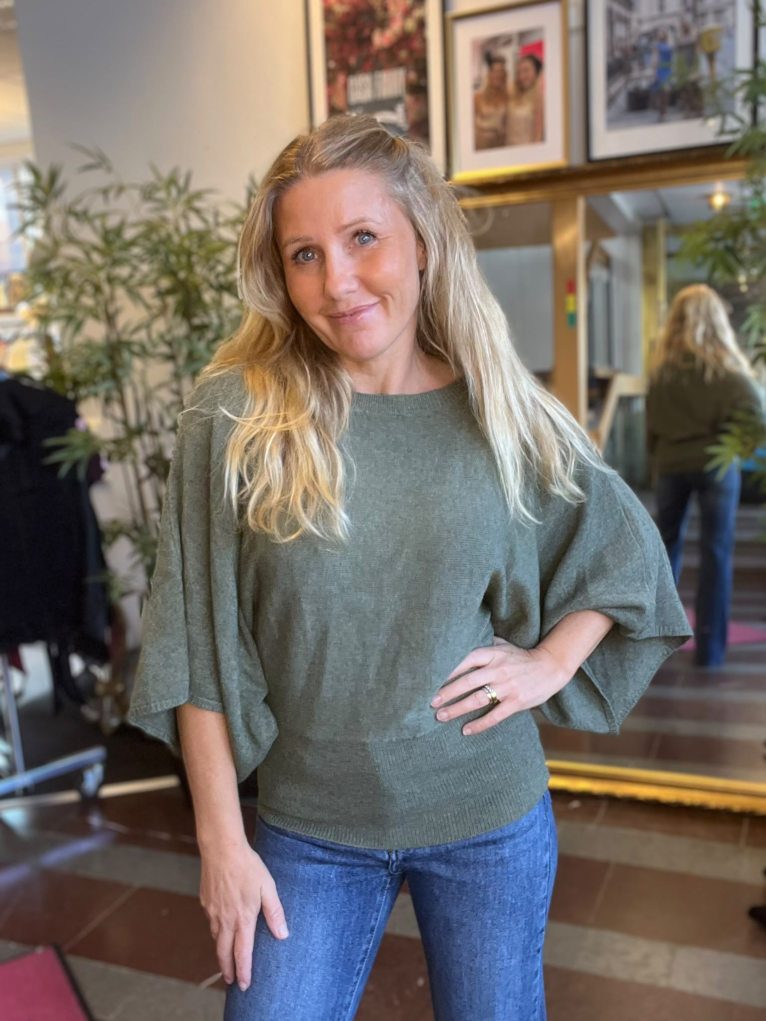 Blond kvinna i grön stickad tröja och blå jeans står och ler mot kameran med handen i sidan. I bakgrunden syns krukväxter, en spegel och inramade foton i ett inomhusrum.