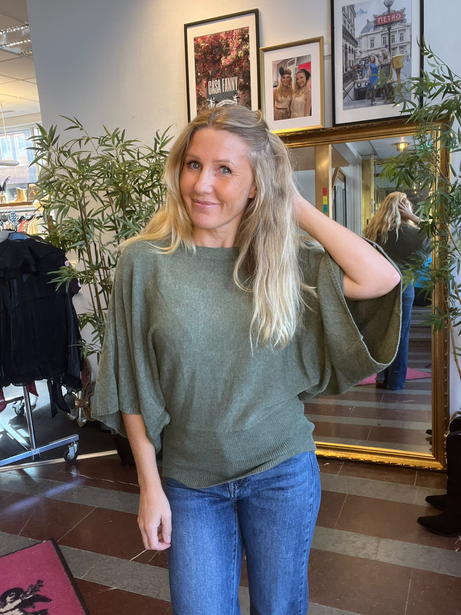 Leende kvinna med långt blont hår står inomhus och håller ena handen bakom huvudet. Hon bär grön stickad topp och jeans; bakom syns spegel, gröna växter och inramade foton med texten “CASA FANNY” och “METRO”.