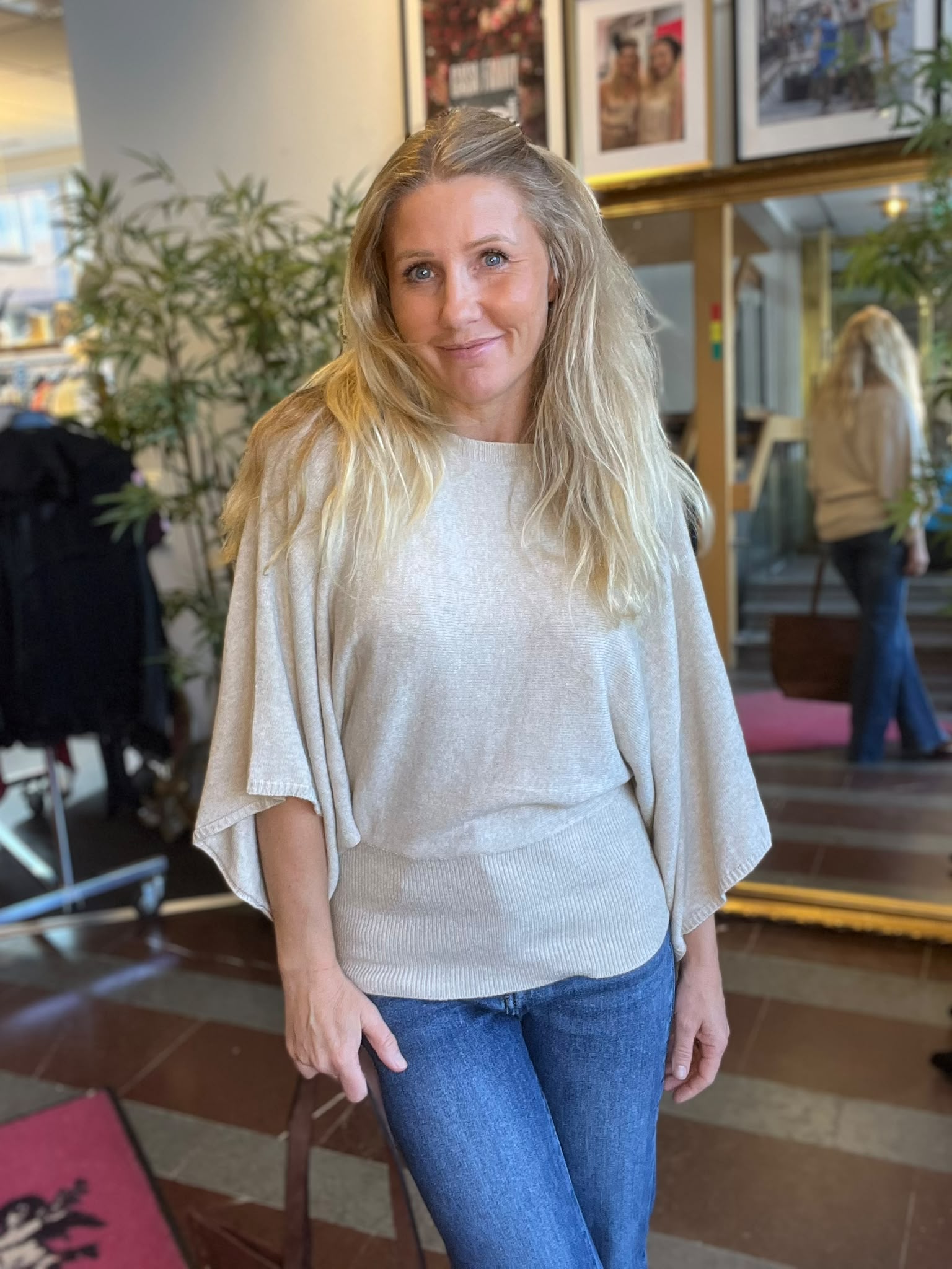 Leende kvinna med långt blont hår står och tittar mot kameran i en ljus inomhusmiljö, klädd i beige stickad topp och blå jeans. Bakom syns växter, inramade foton och en spegel.