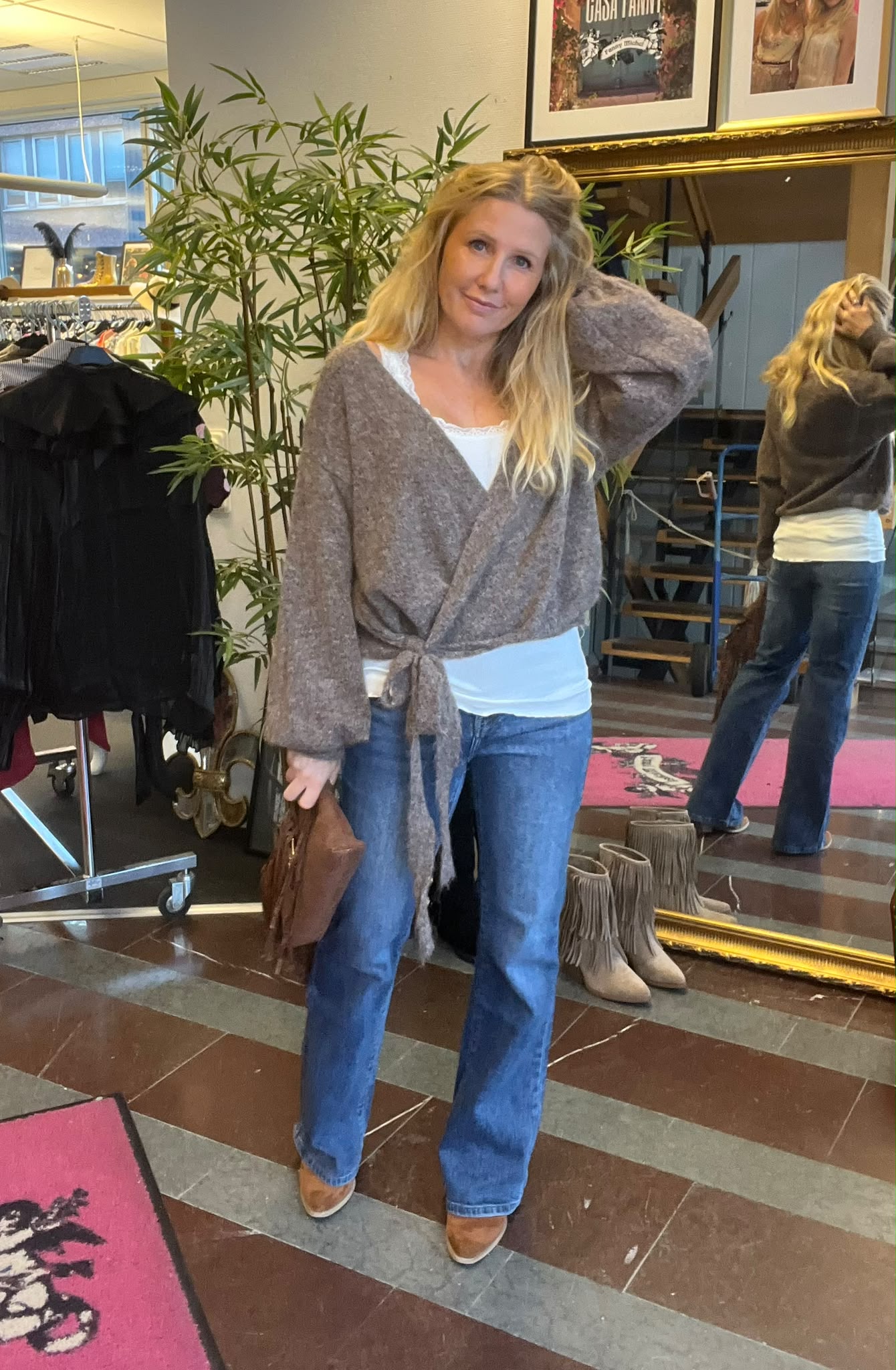 Blond kvinna poserar i en klädbutik, med ena handen i håret och den andra som håller en brun väska. Hon bär grå omlottkofta, vit topp och blå jeans, framför en stor spegel och klädställning.