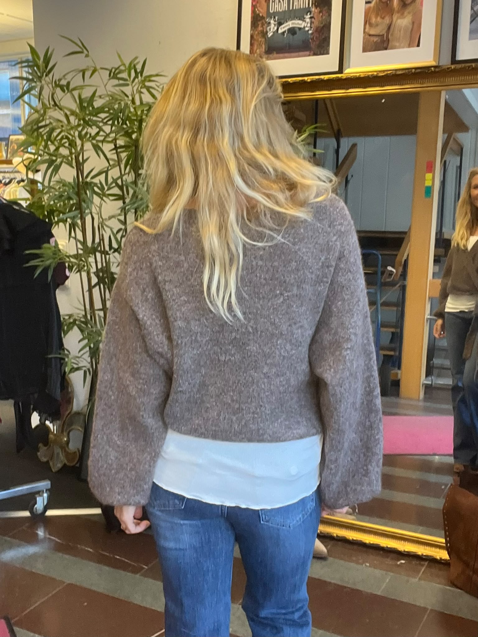 Kvinna med långt blont hår står med ryggen mot kameran och tittar i en stor guldramad spegel. Hon bär grå stickad tröja och jeans. I rummet syns en grön växt, tavlor och hennes spegelbild.