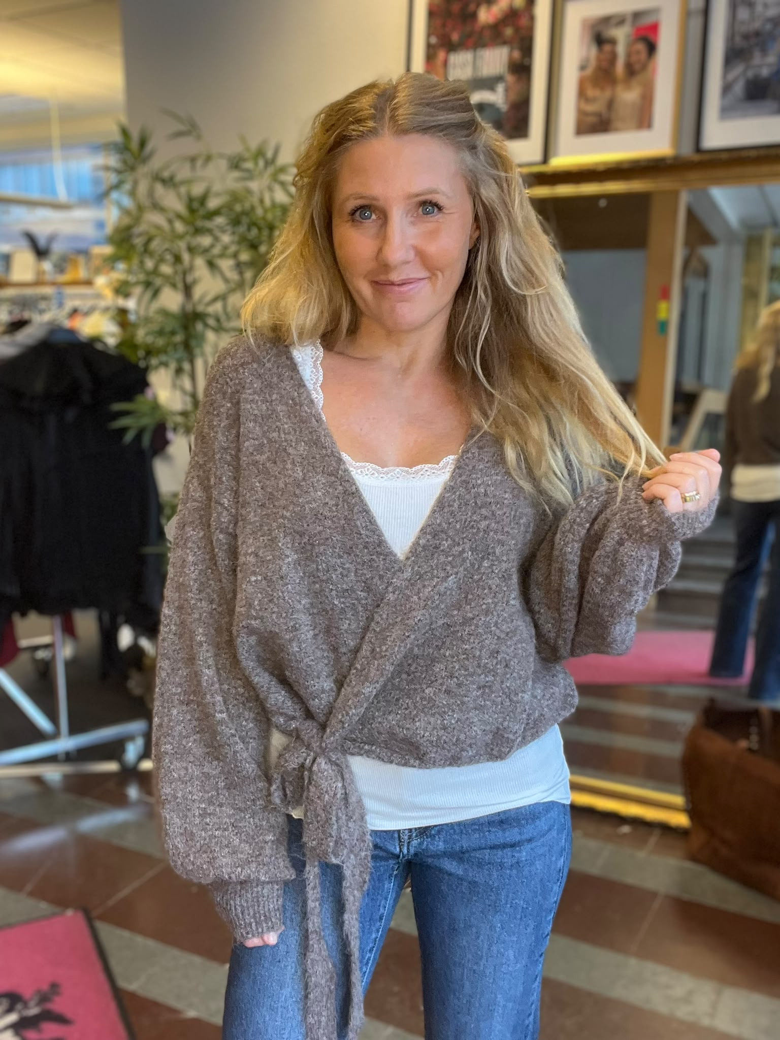Leende kvinna med långt blont hår poserar inomhus och håller en hårslinga i handen. Hon bär grå stickad omlottkofta över vitt linne och jeans; bakom syns spegel, växt, klädställning och inramade foton.