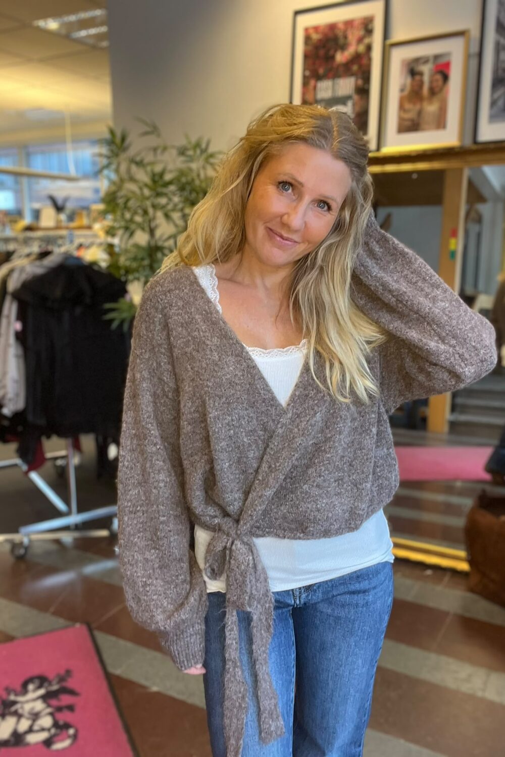 Leende kvinna med långt blont hår står i en butiksmiljö och håller ena handen bakom huvudet. Hon bär en brun stickad omlottkofta över vit topp och jeans; bakom syns klädställningar, växter och inramade foton.