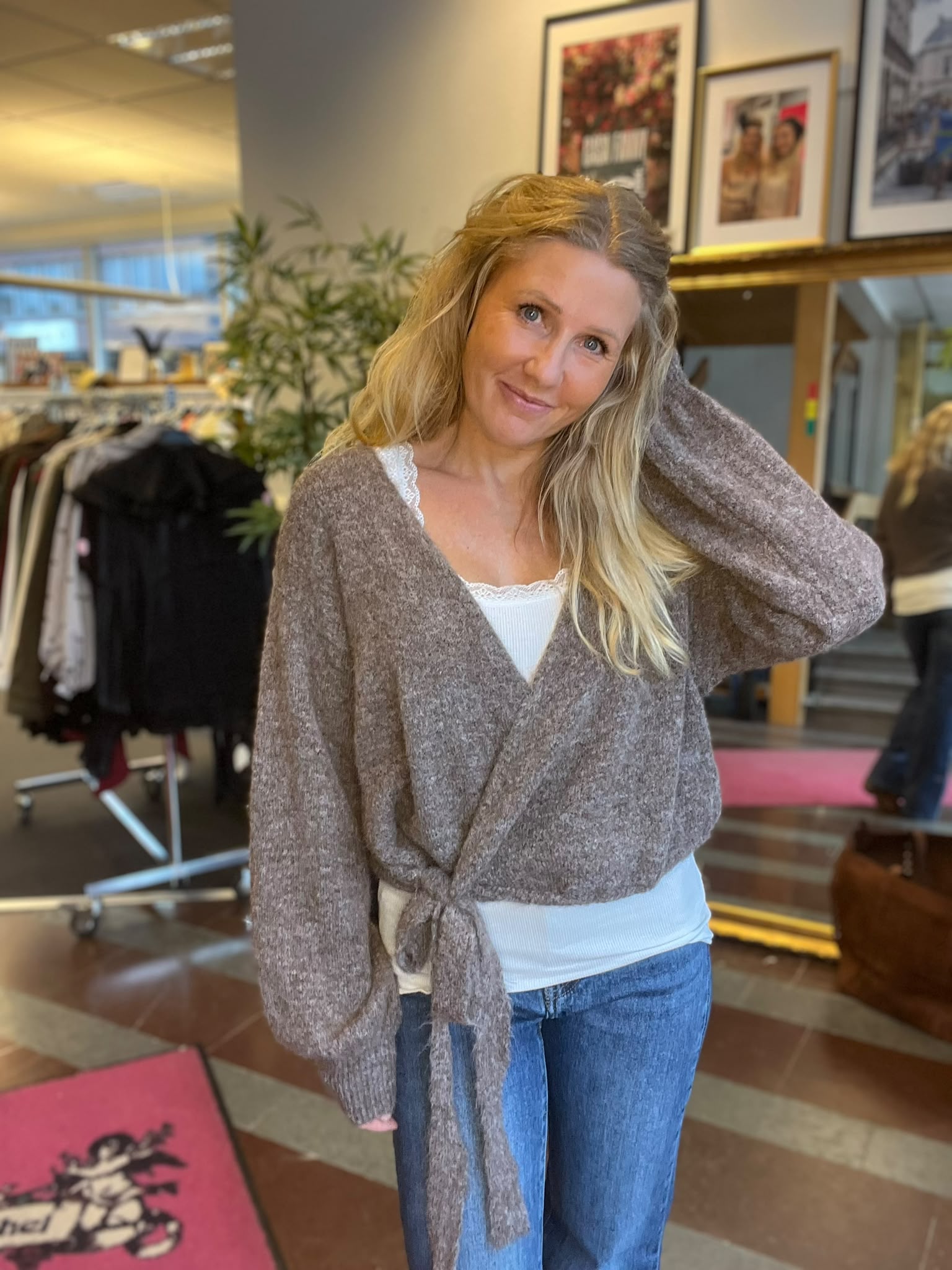 Leende kvinna med långt blont hår står i en butiksmiljö och håller ena handen bakom huvudet. Hon bär en brun stickad omlottkofta över vit topp och jeans; bakom syns klädställningar, växter och inramade foton.