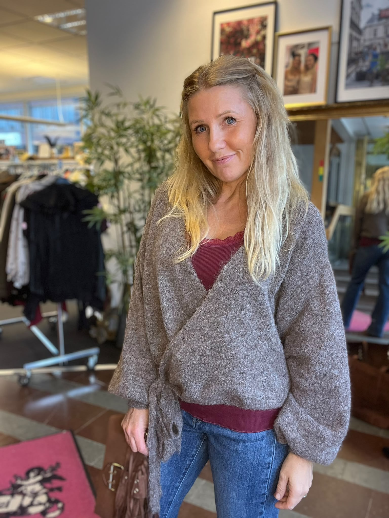 Kvinna med långt blont hår står och ler mot kameran i en inomhusmiljö, iklädd grå kofta, vinröd topp och jeans. I bakgrunden syns en klädställning, en stor grön växt och inramade foton.