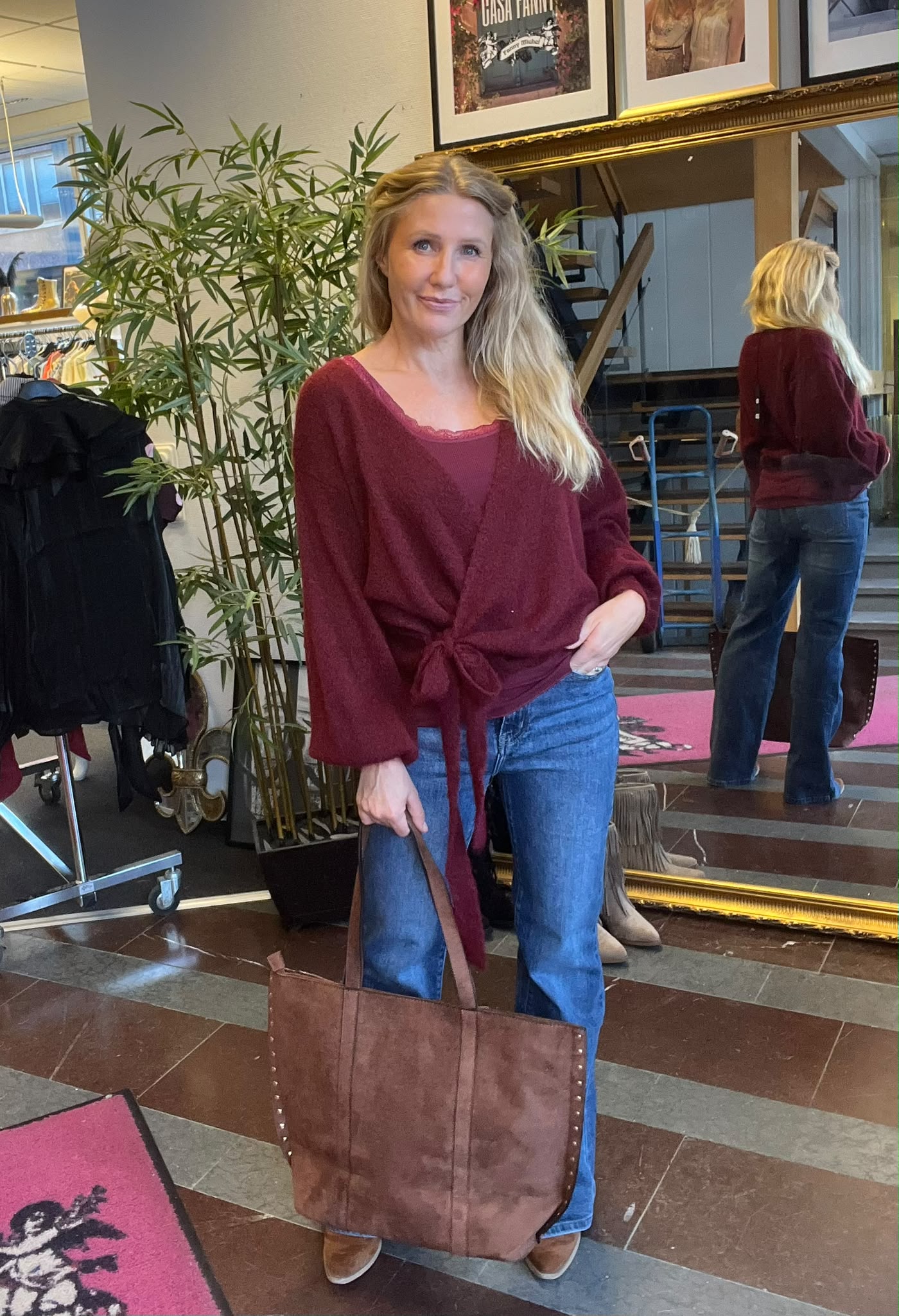 Kvinna med långt blont hår står och poserar med handen i jeansfickan och håller en stor brun läderväska. Hon bär vinröd knuten tröja. I bakgrunden syns spegel, växt och klädställning i butiksmiljö.
