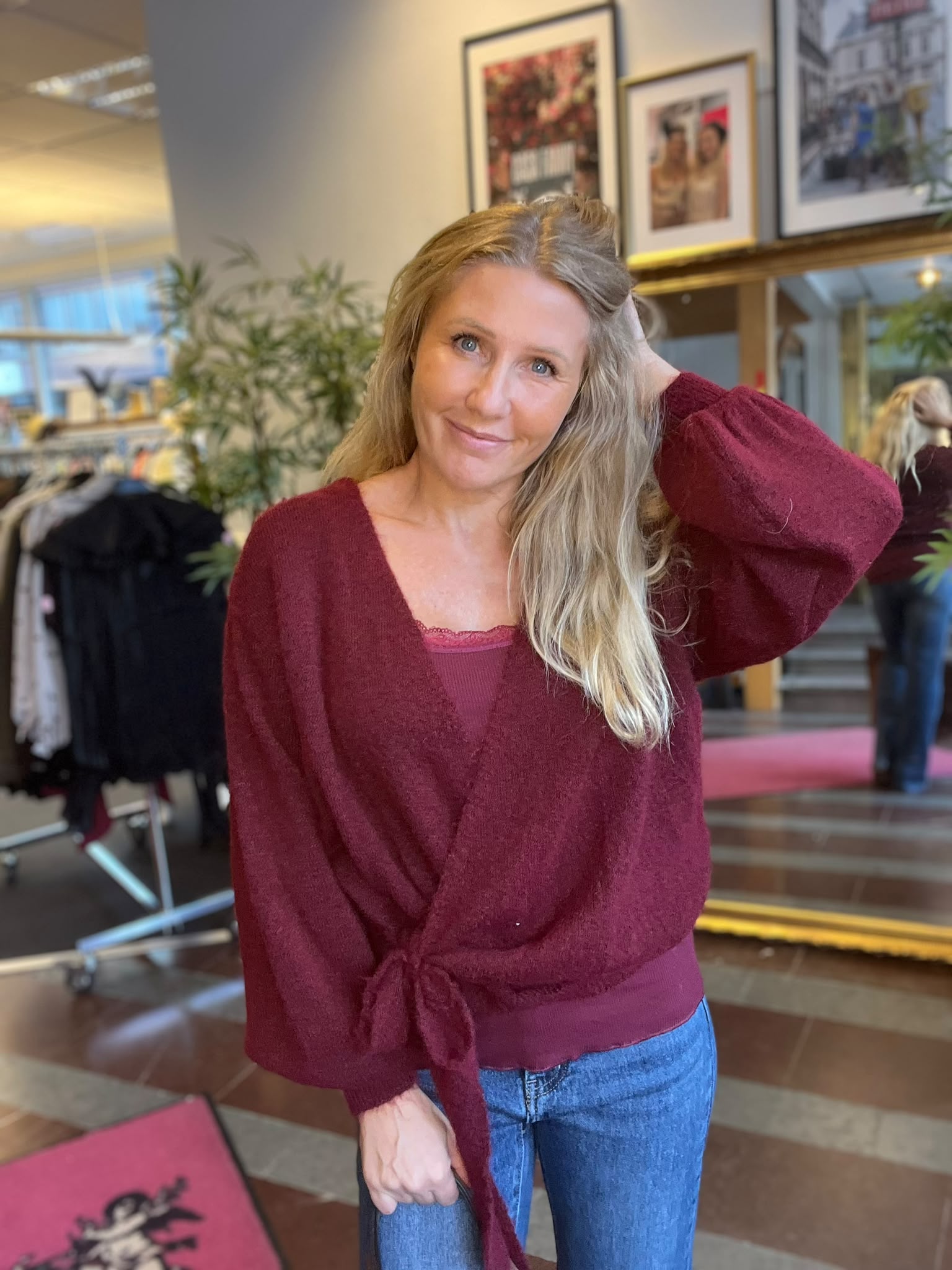 Leende kvinna med långt blont hår poserar med ena handen bakom huvudet och den andra på en stol. Hon bär vinröd stickad tröja och jeans i en ljus lokal med växter och inramade tavlor.