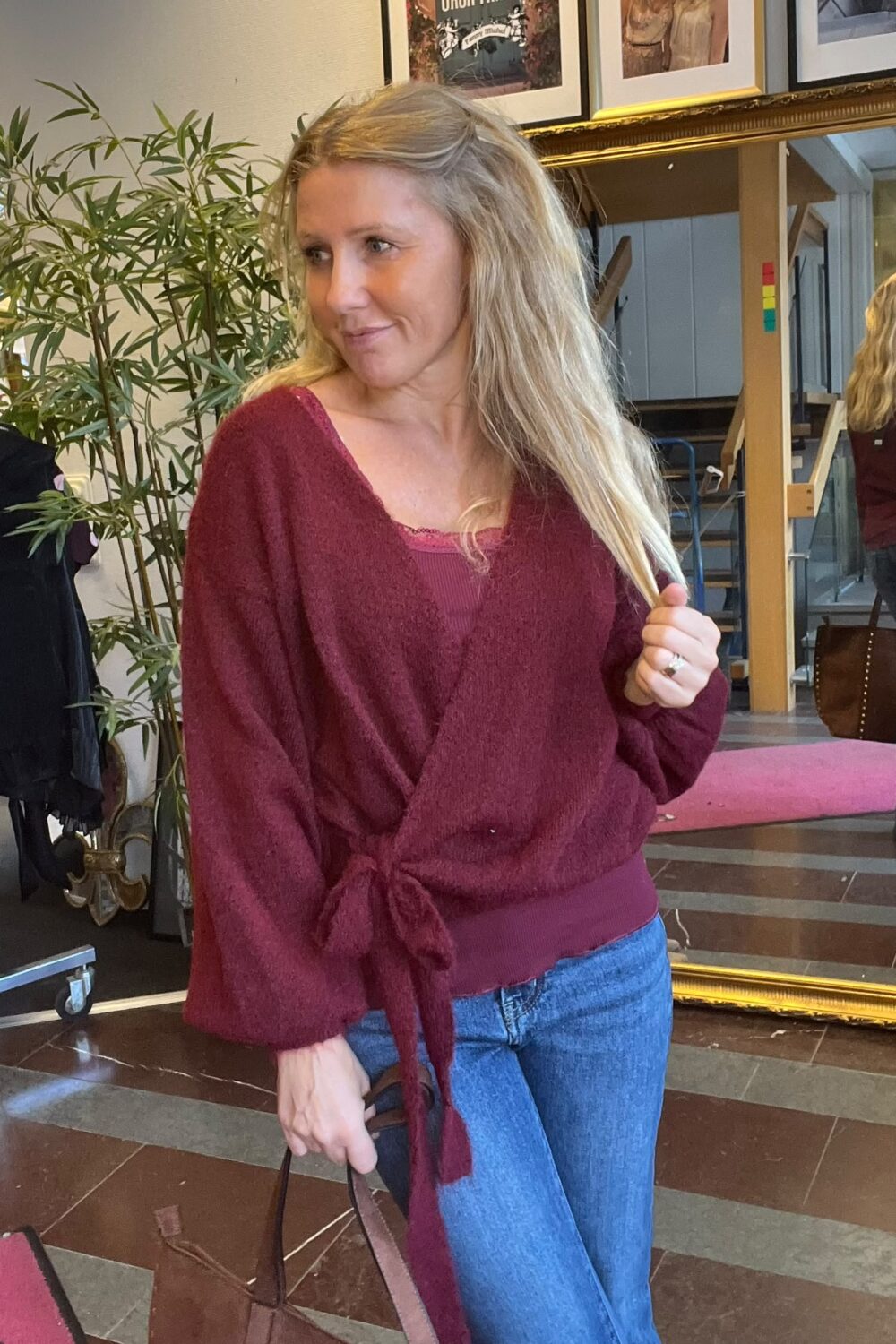 Blond kvinna i vinröd stickad tröja och jeans står inomhus och håller i en handväska medan hon ser åt sidan. Bakom henne syns stor spegel, grön växt och inramade tavlor på väggen.