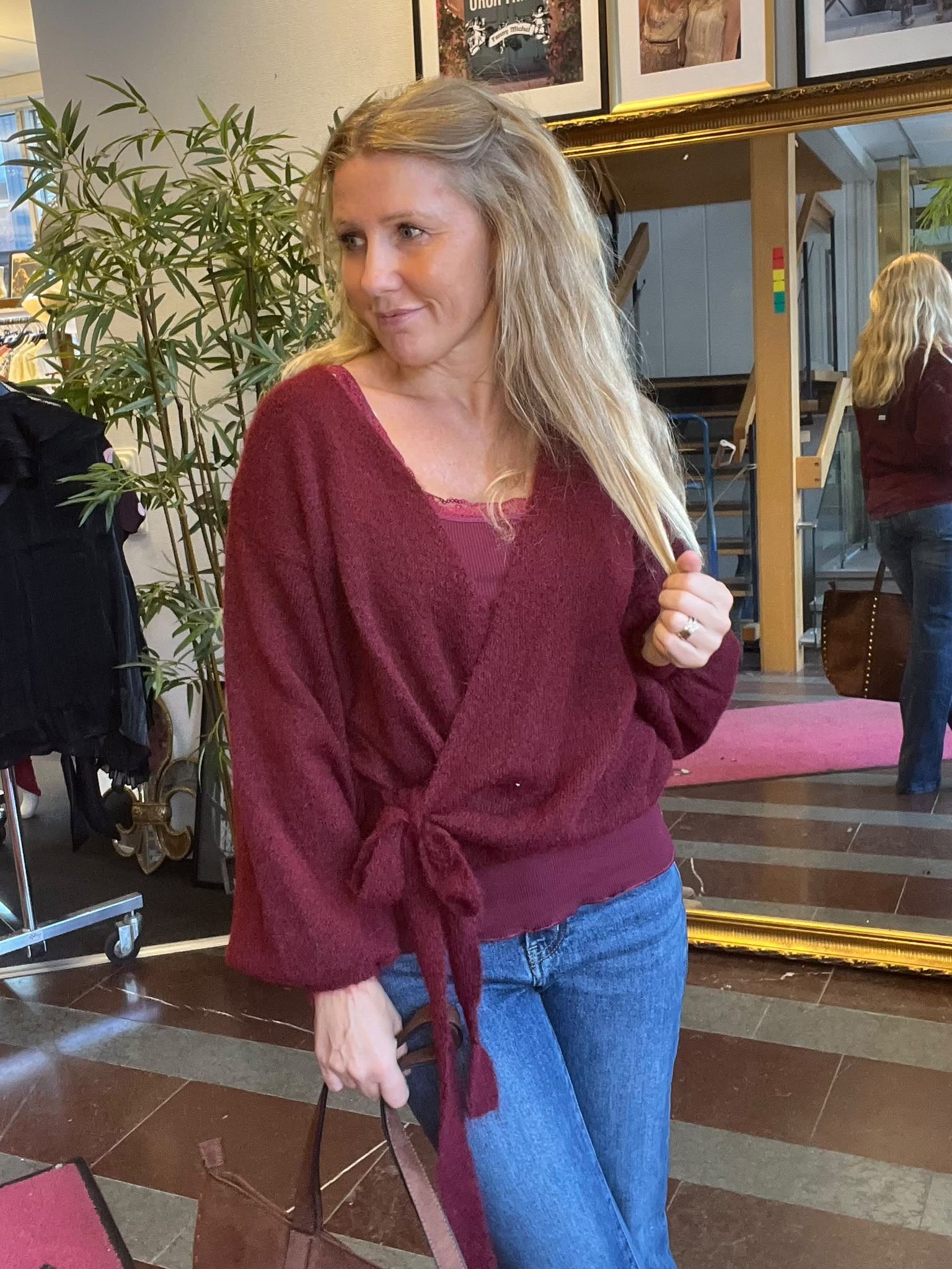 Blond kvinna i vinröd stickad tröja och jeans står inomhus och håller i en handväska medan hon ser åt sidan. Bakom henne syns stor spegel, grön växt och inramade tavlor på väggen.