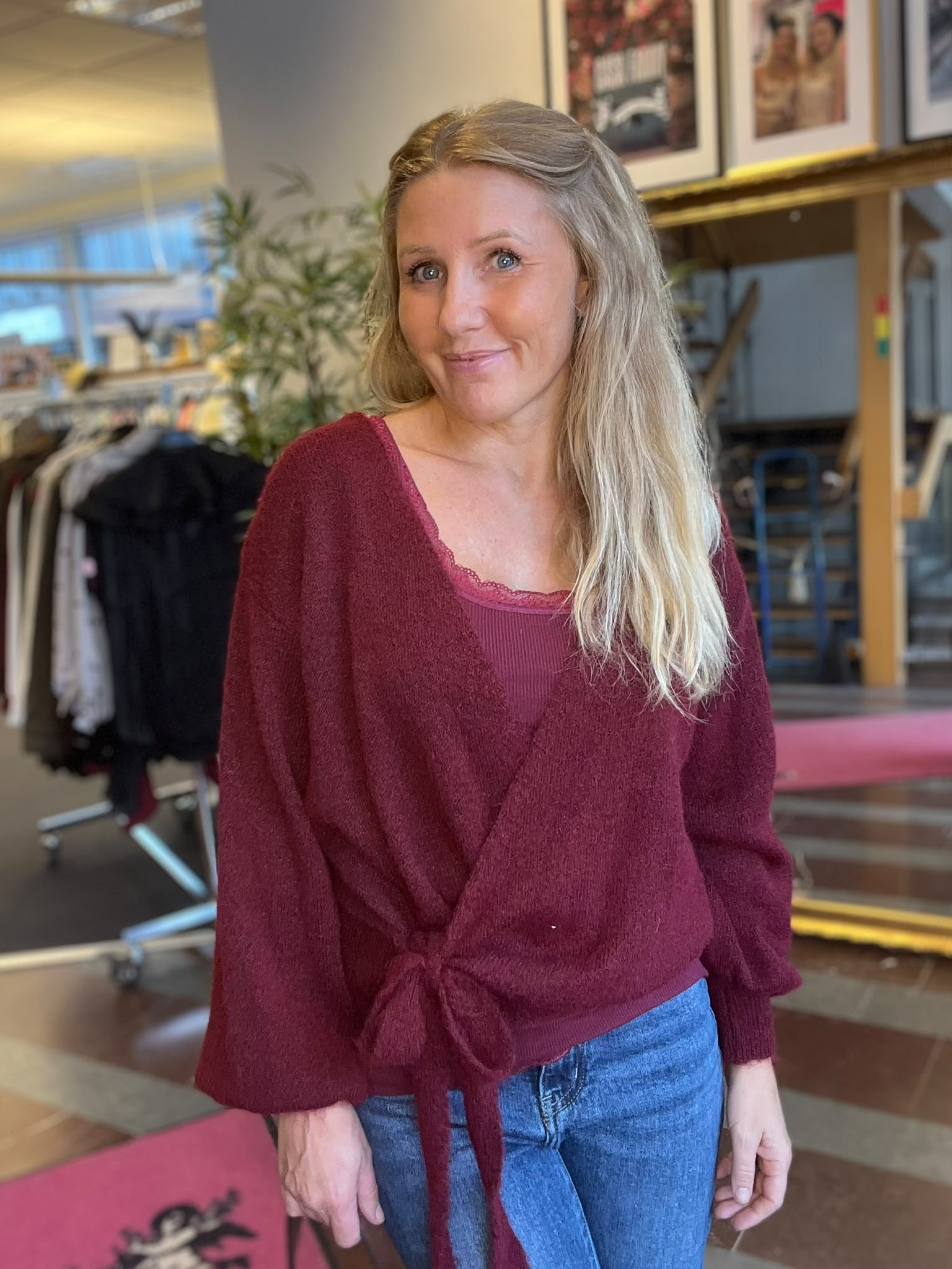 Leende kvinna med långt blont hår står och poserar i en vinröd omlotttröja och jeans. Hon tittar mot kameran i en inomhusmiljö med klädställning, växt och inramade foton i bakgrunden.