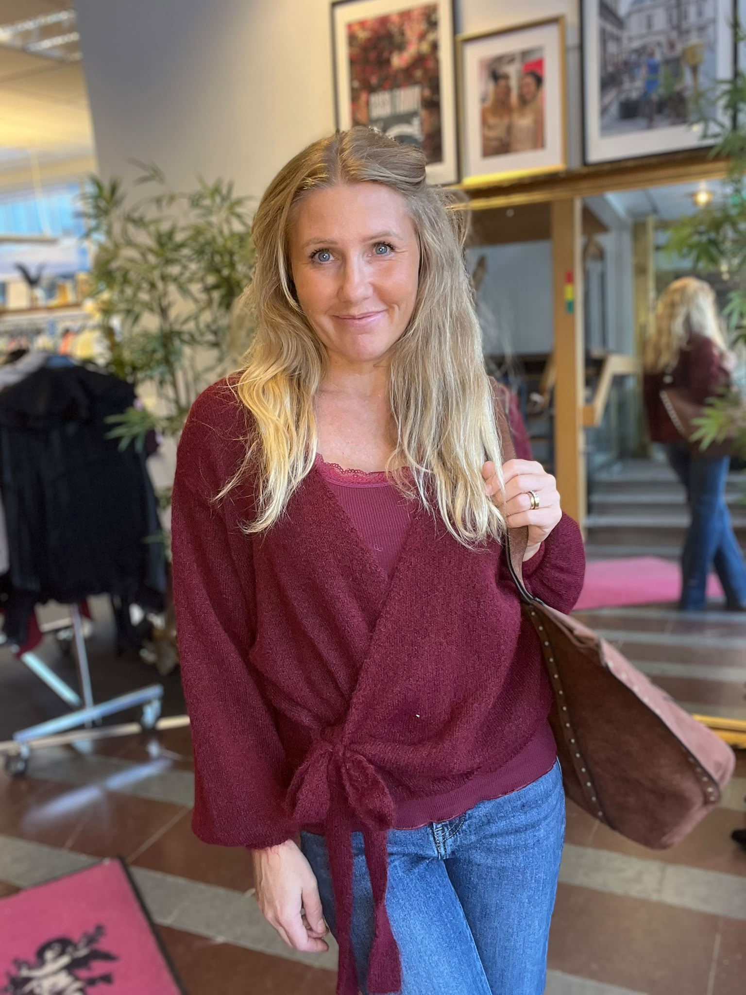 Leende kvinna med långt blont hår står inomhus och håller i remmen till en brun axelväska. Hon bär vinröd kofta och jeans. I bakgrunden syns växter, inramade foton och en spegel.