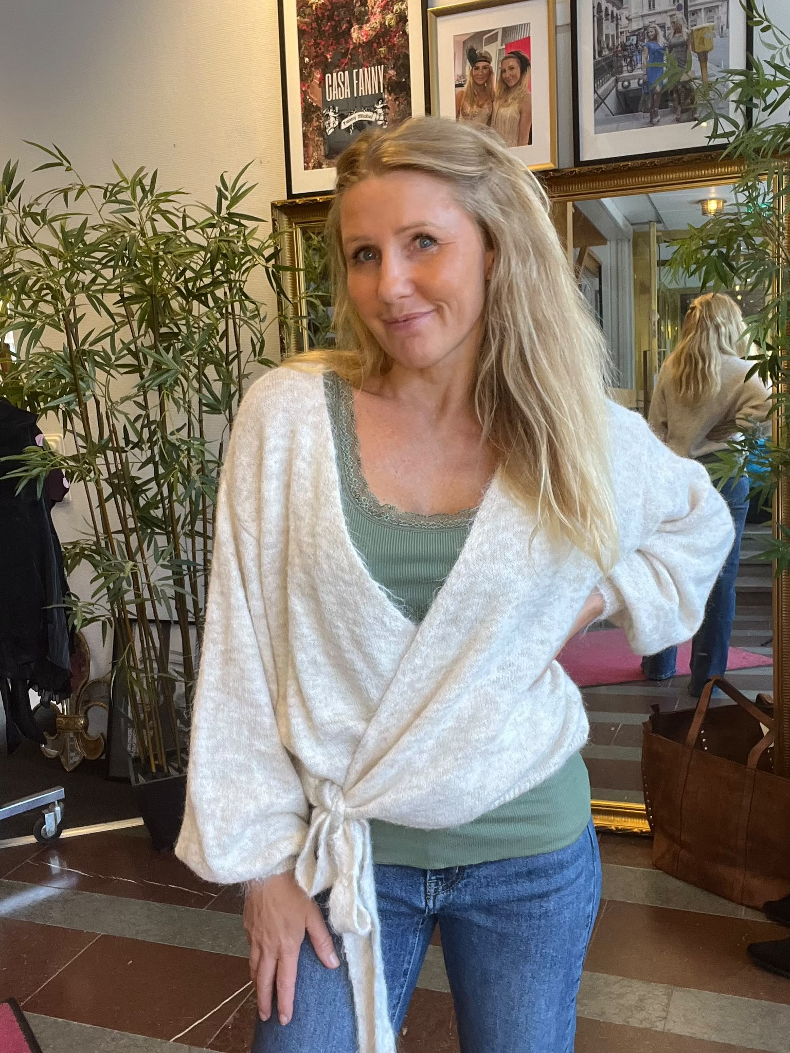 Leende kvinna med långt blont hår står med handen på höften i en inomhusmiljö, klädd i grön topp, jeans och vit knuten kofta. Bakom syns spegel, växter och inramade bilder med texten “CASA FANNY”.
