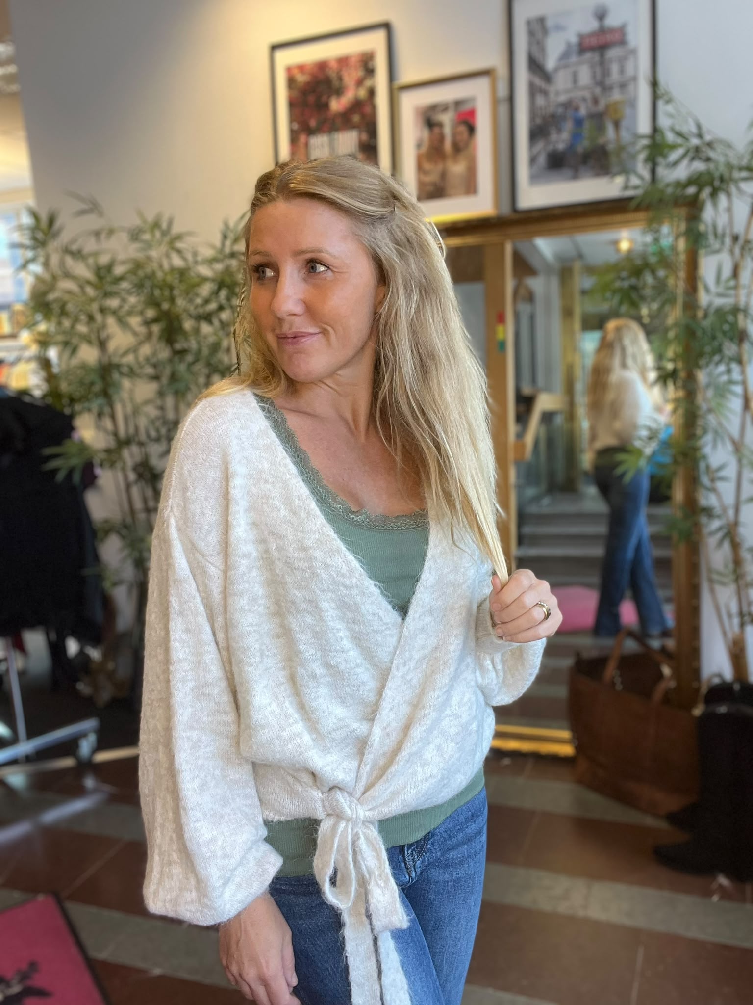 Blond kvinna står inomhus och tittar åt sidan medan hon håller i sin ljusa kofta över ett grönt linne och jeans. I bakgrunden syns växter, inramade tavlor och en spegel som reflekterar rummet.