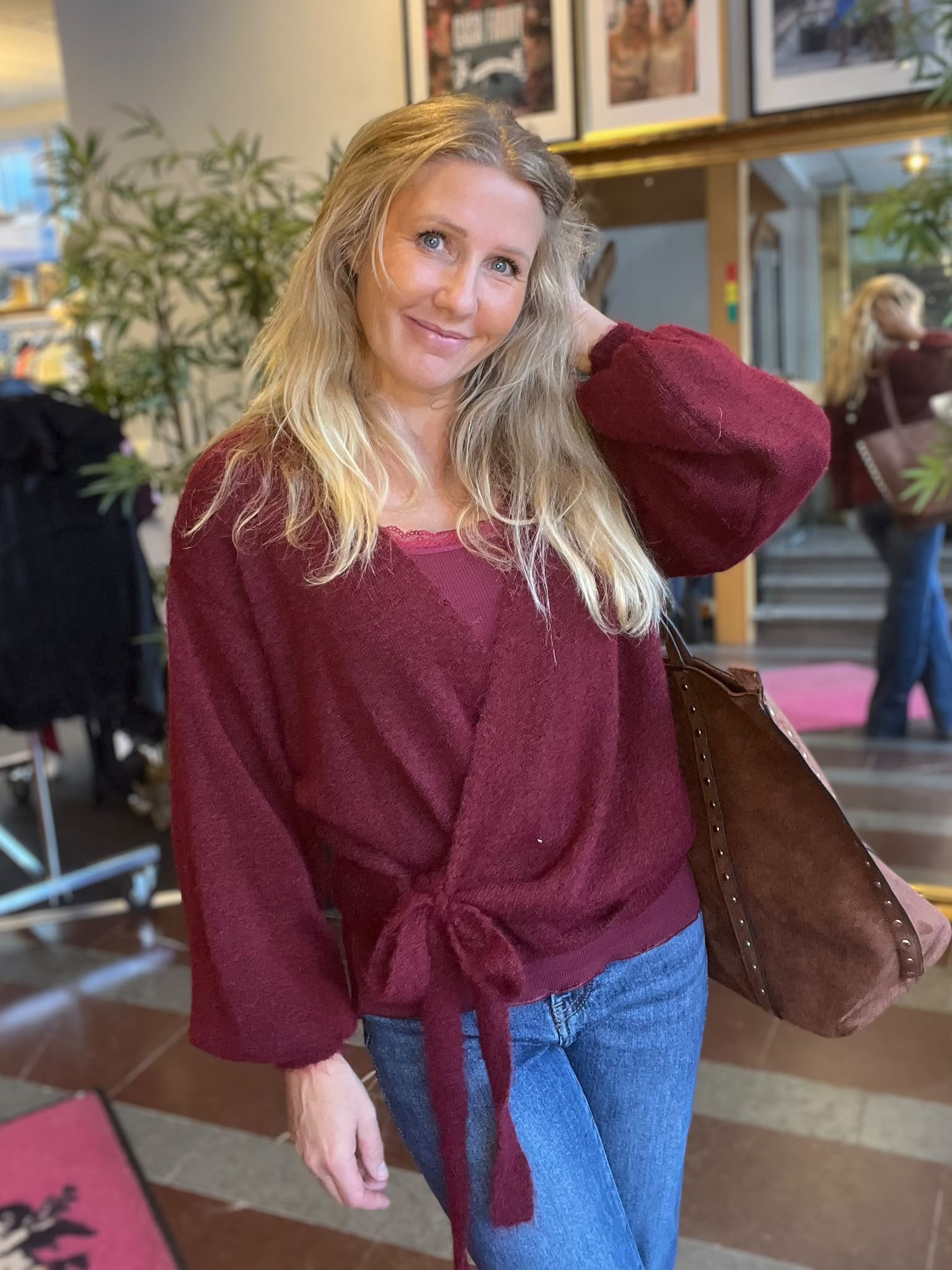 Leende blond kvinna i vinröd stickad tröja och jeans poserar med ena handen i håret och en brun axelväska. Hon står inomhus i en lobby med växter, tavlor och en spegel i bakgrunden.