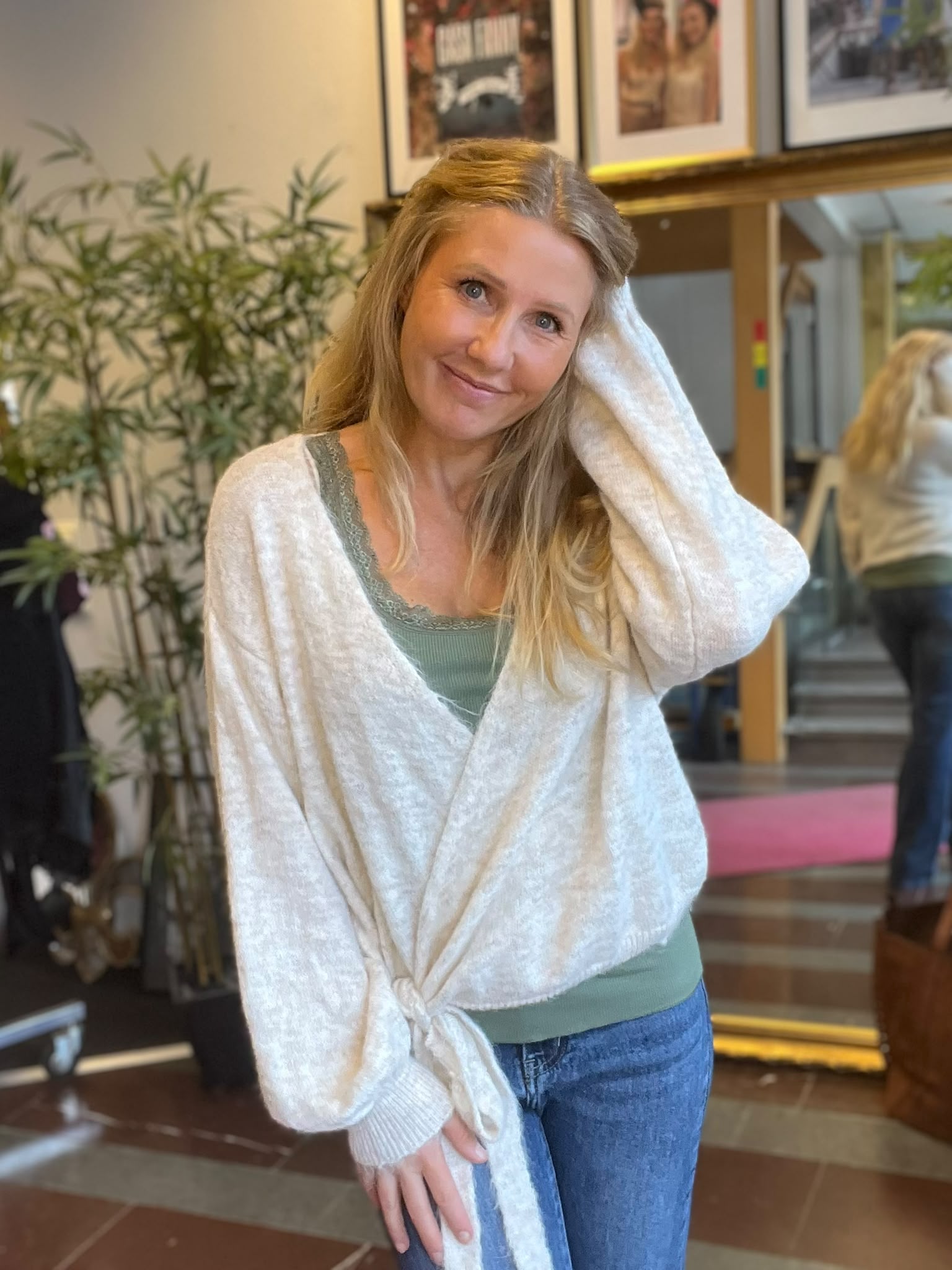 Leende blond kvinna lutar huvudet mot sin hand och tittar mot kameran, klädd i vit kofta, grön topp och jeans. Hon står inomhus vid en stor spegel, växt och inramade foton i bakgrunden.