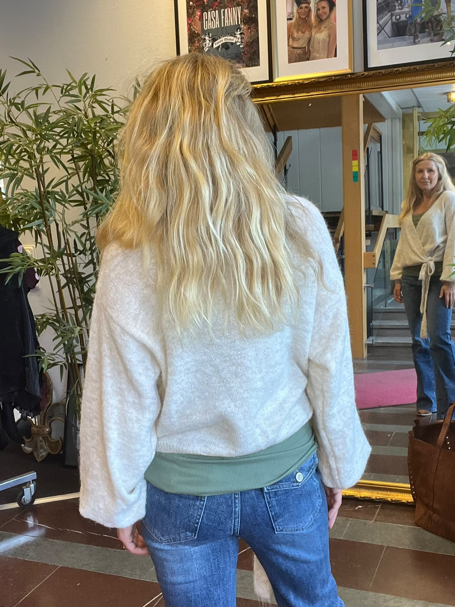 Kvinna med långt blont hår står med ryggen mot kameran och tittar på sin spegelbild i en stor guldram. Inomhusmiljö med växter och inramade bilder ovanför, inklusive texten “CASA FANNY”.