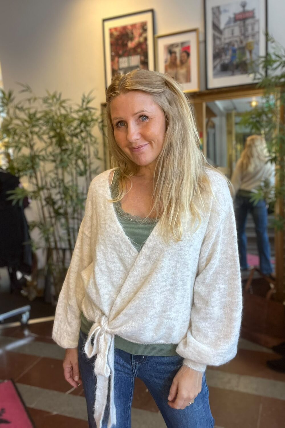Blond kvinna ler mot kameran och står avslappnat med armarna vid sidan, klädd i grön topp, jeans och ljus omlottkofta. Inomhusmiljö med växter, spegel och inramade tavlor i bakgrunden.