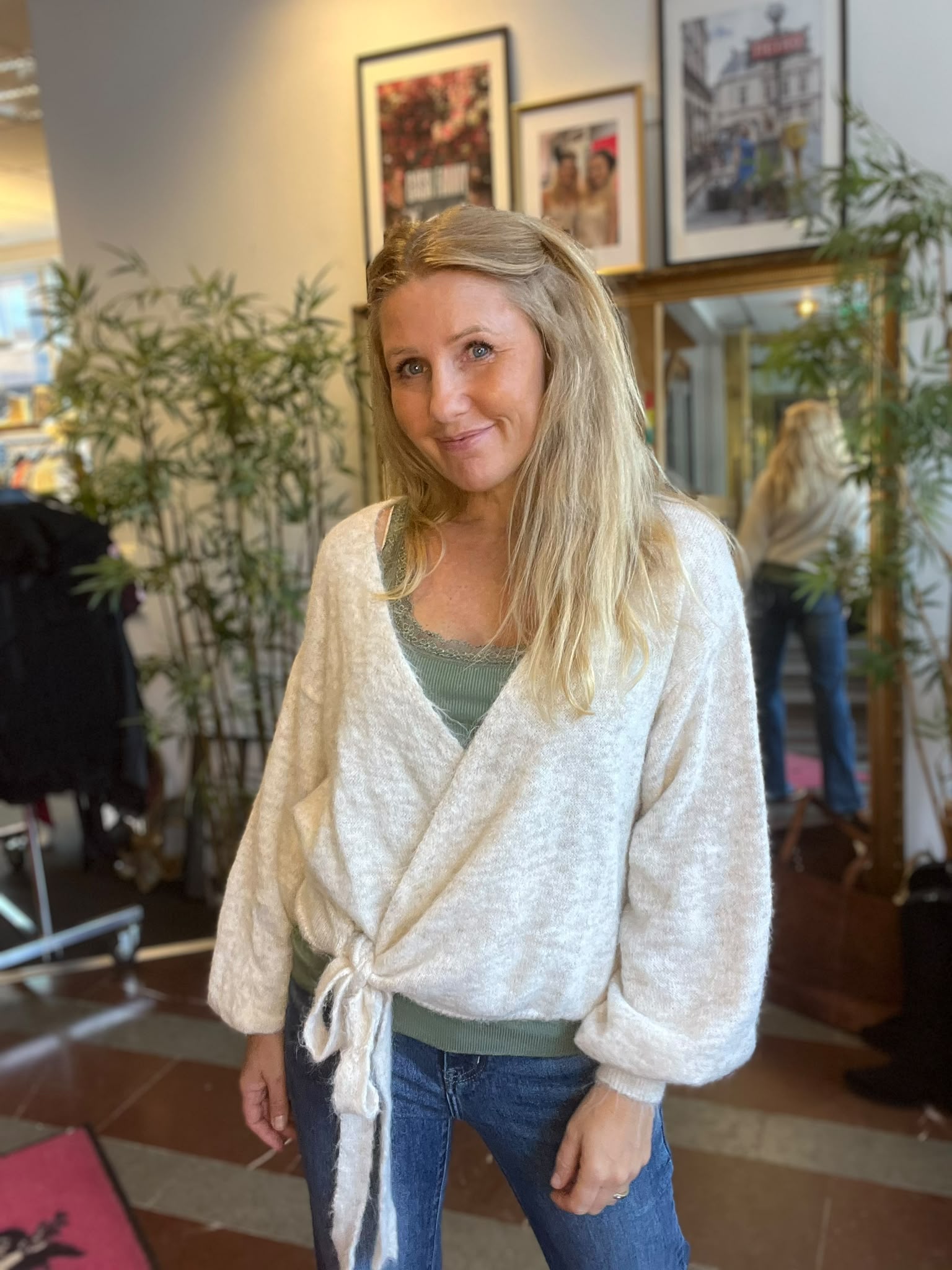 Blond kvinna ler mot kameran och står avslappnat med armarna vid sidan, klädd i grön topp, jeans och ljus omlottkofta. Inomhusmiljö med växter, spegel och inramade tavlor i bakgrunden.
